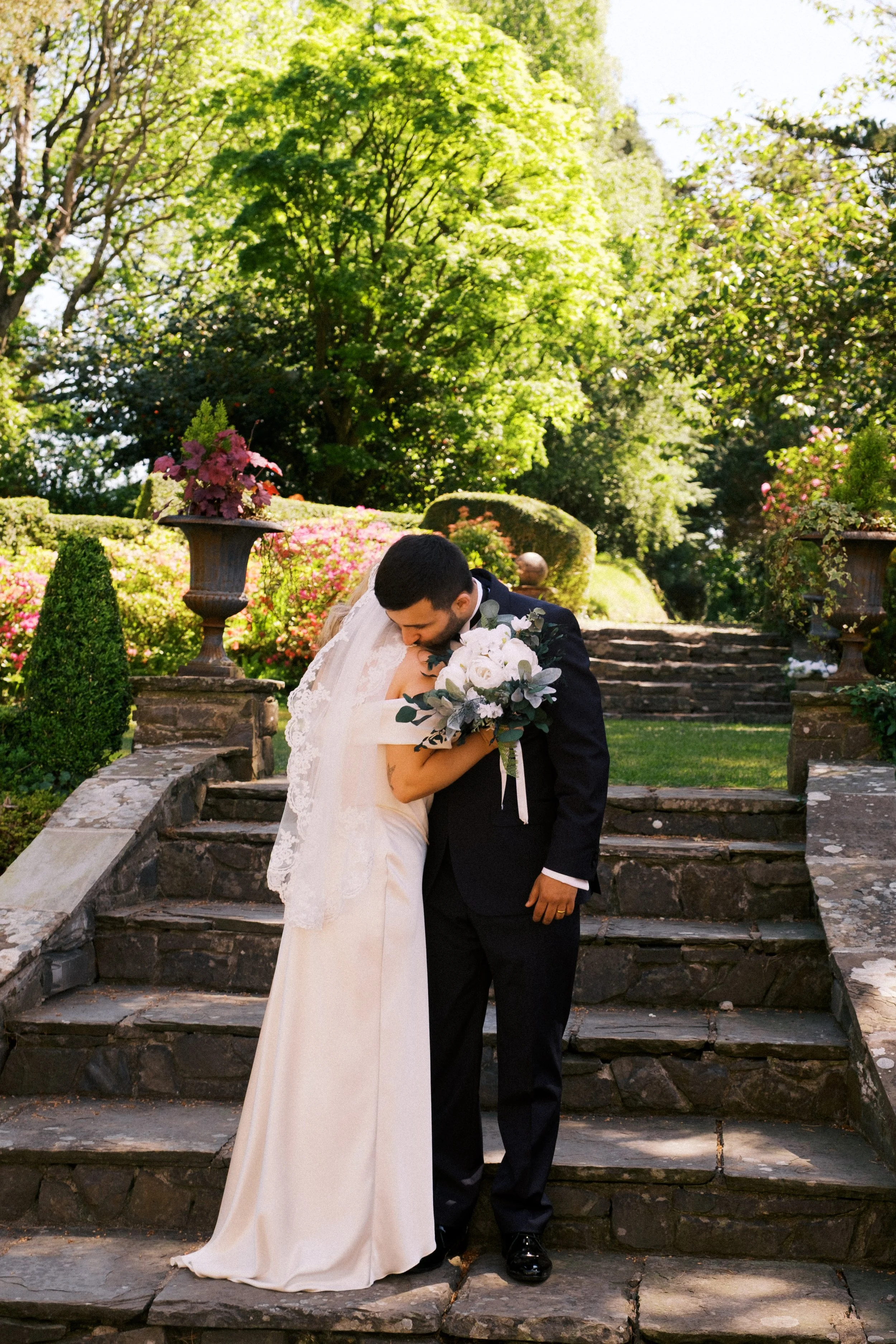 A bride and groom sharing a kiss on stone steps in a garden with lush green trees, pink flowers in urns, and sunlight filtering through the leaves.