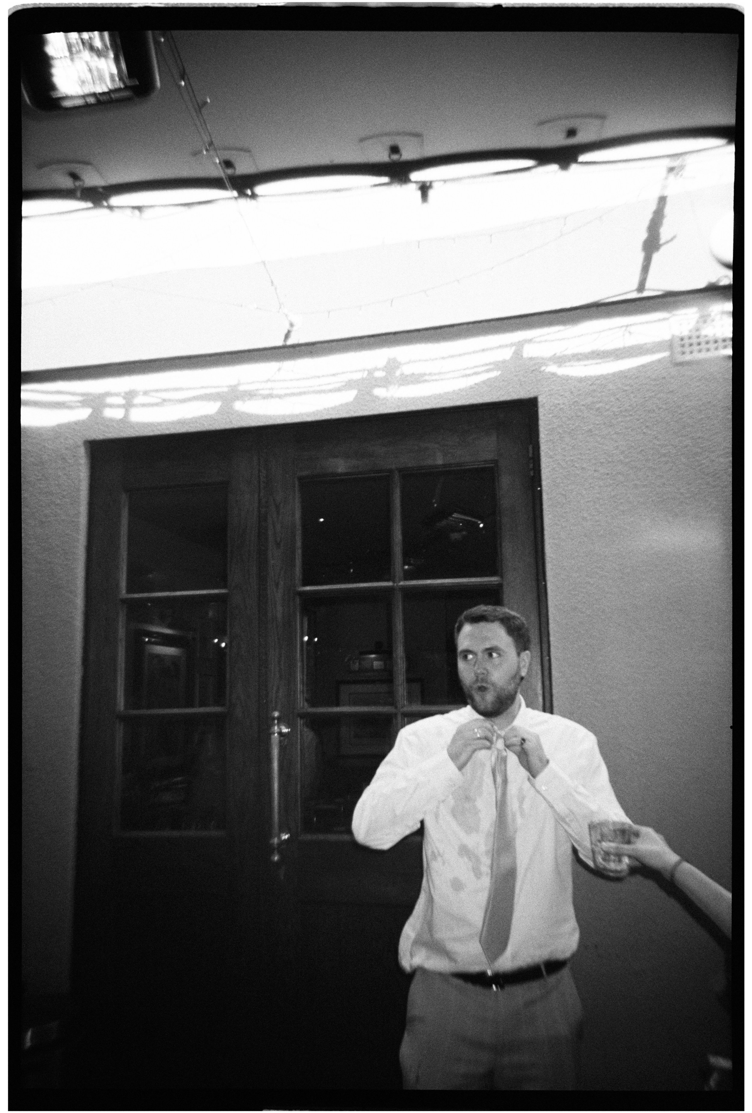 Black and white photo of a man in a dress shirt and tie holding a glass of drink, with someone else partially visible holding his tie, inside a room with a window and a wood door in the background.