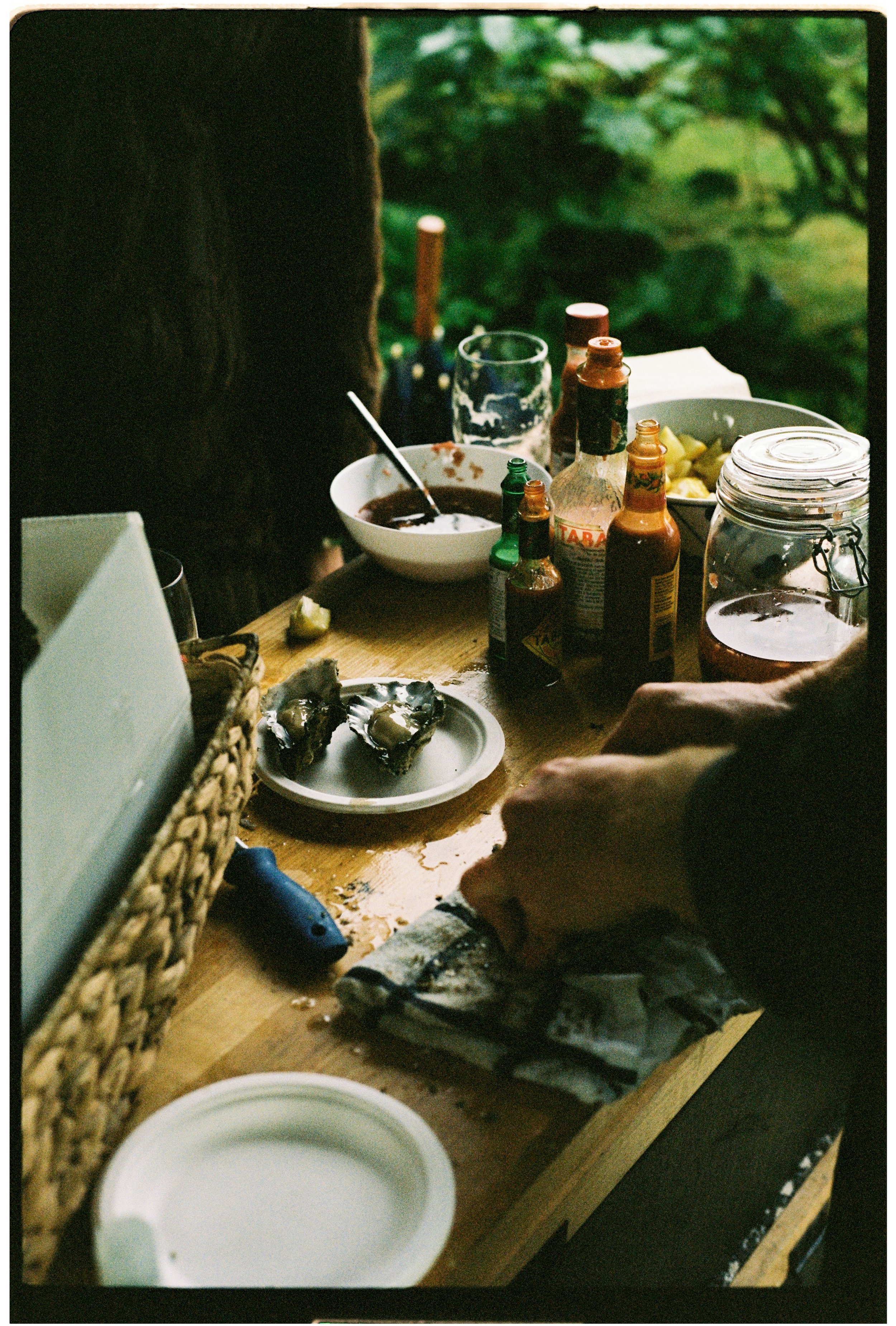 A table with various condiments, a bowl of sauce, a jar with a lid, a glass container, and some food items. A person's hand is seen holding a spoon. The table is outdoors with greenery in the background.