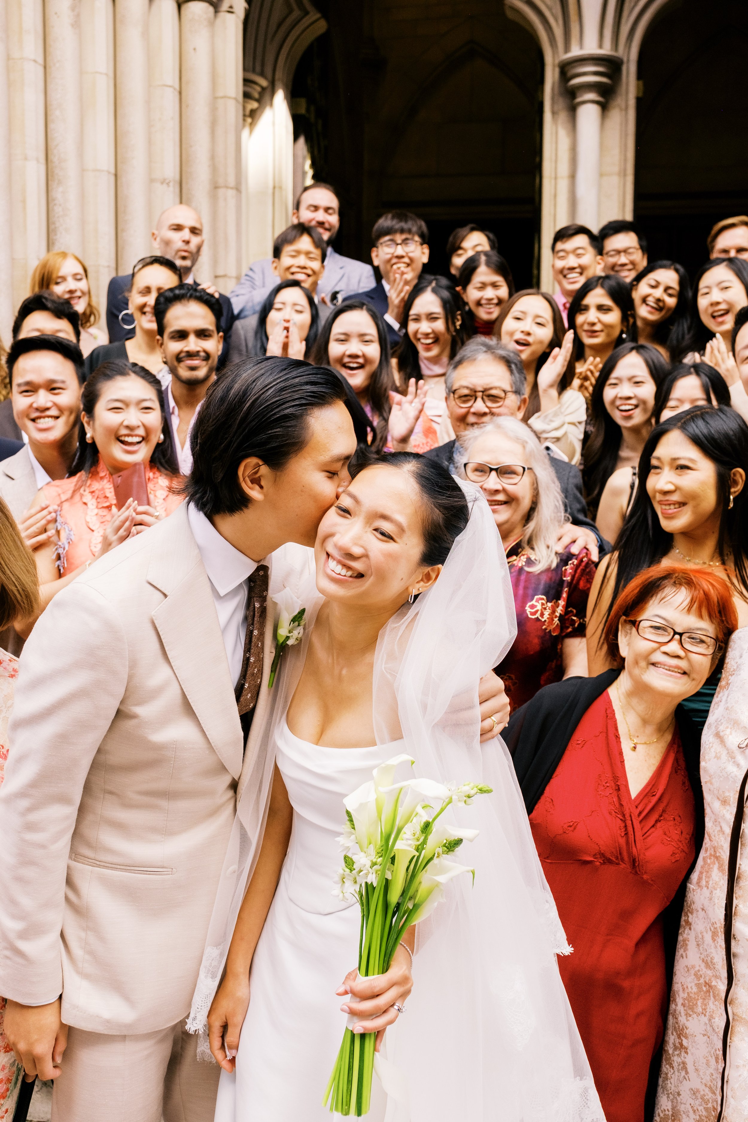 A wedding couple sharing a kiss in front of a large group of smiling friends and family inside a church with tall stone columns.