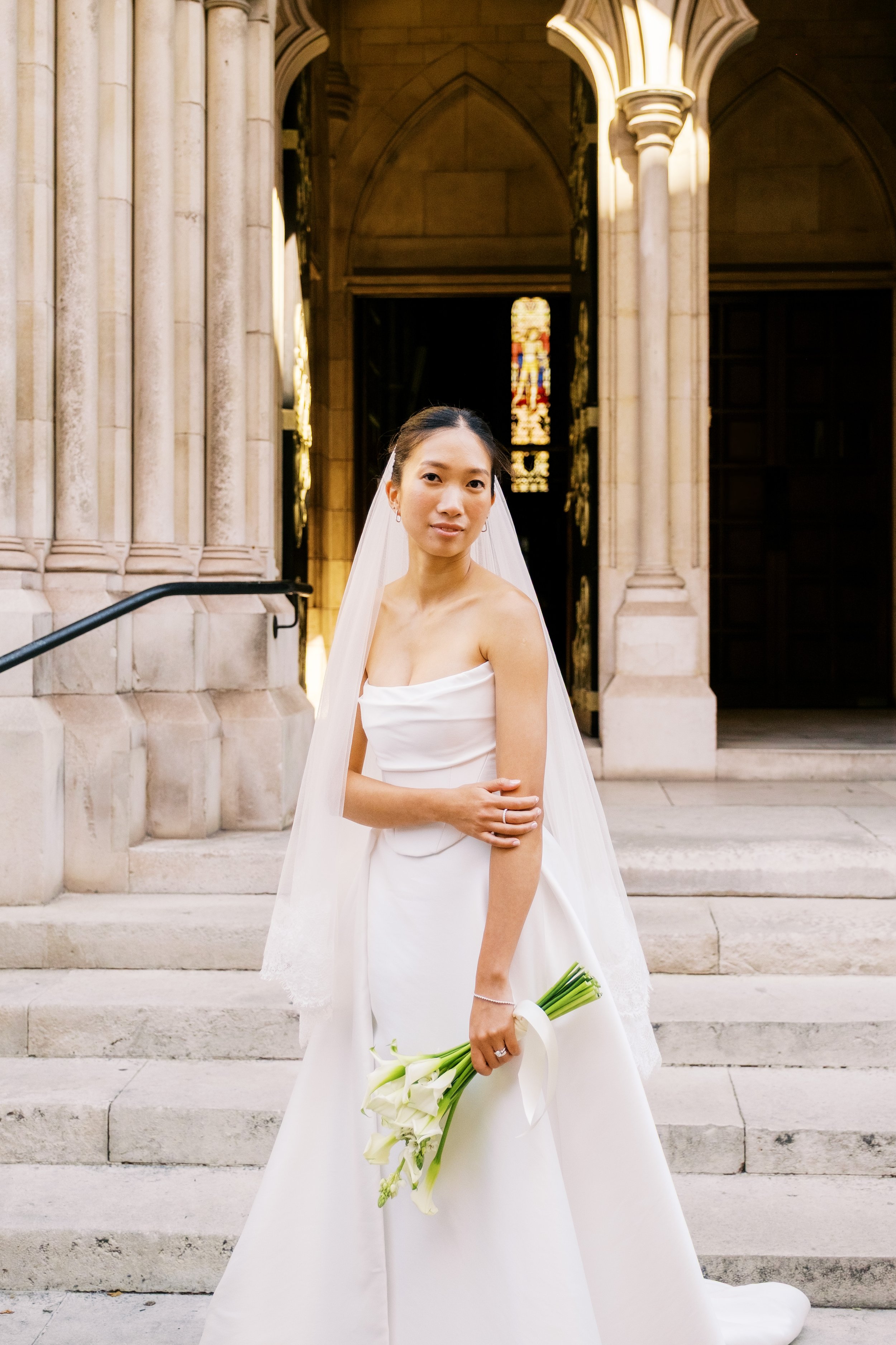 A bride in a white wedding dress holding a bouquet of white calla lilies, standing on stone steps outside a church with Gothic architecture and stained glass windows.