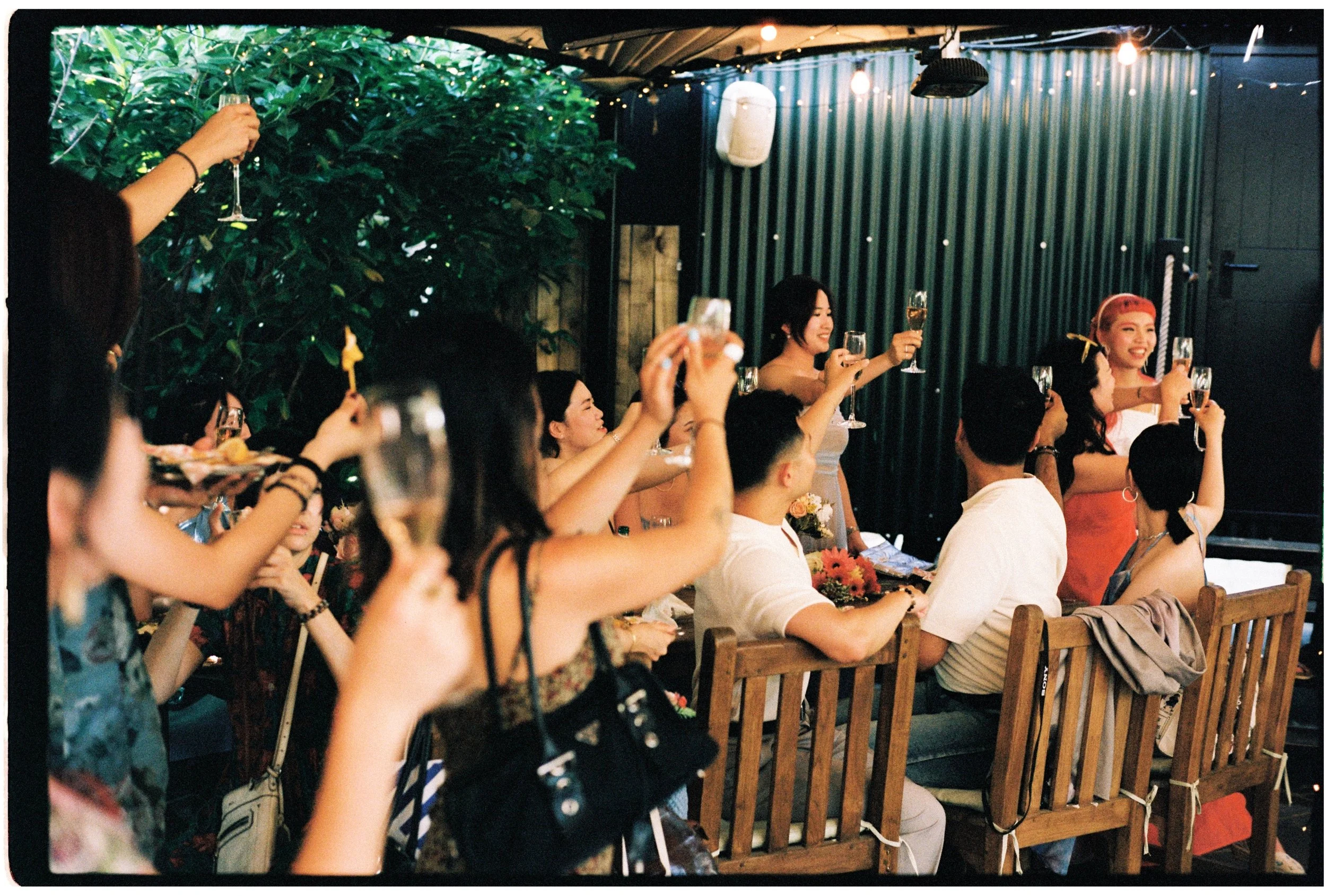 People gathered at a celebration holding up glasses, some sitting on wooden benches, under a covered outdoor area decorated with string lights and greenery.