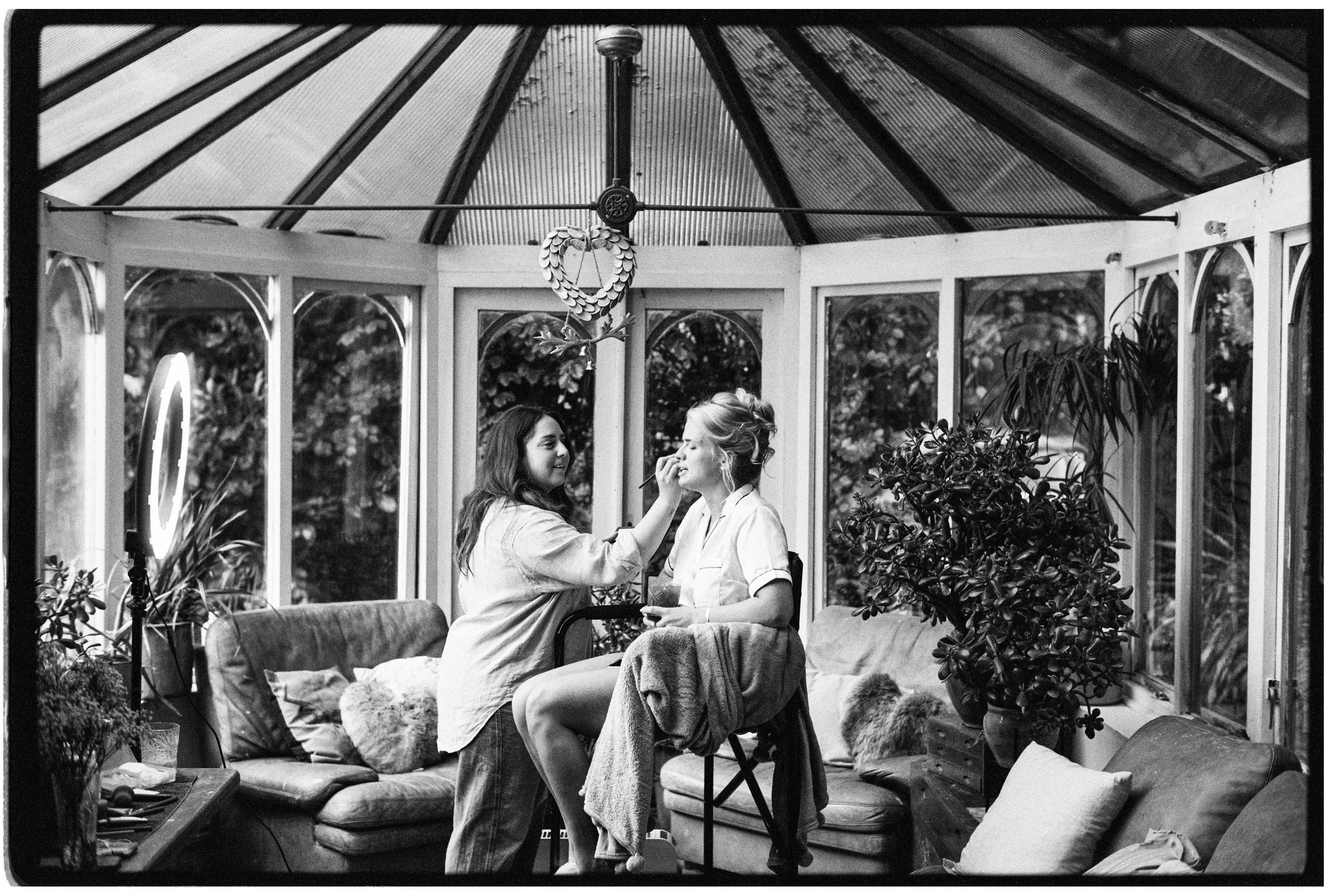 Two women are sitting inside a glass conservatory, one applying makeup to the other. The space is decorated with plants and cushions, and has a corrugated metal roof. There is a mirror with makeup tools nearby.