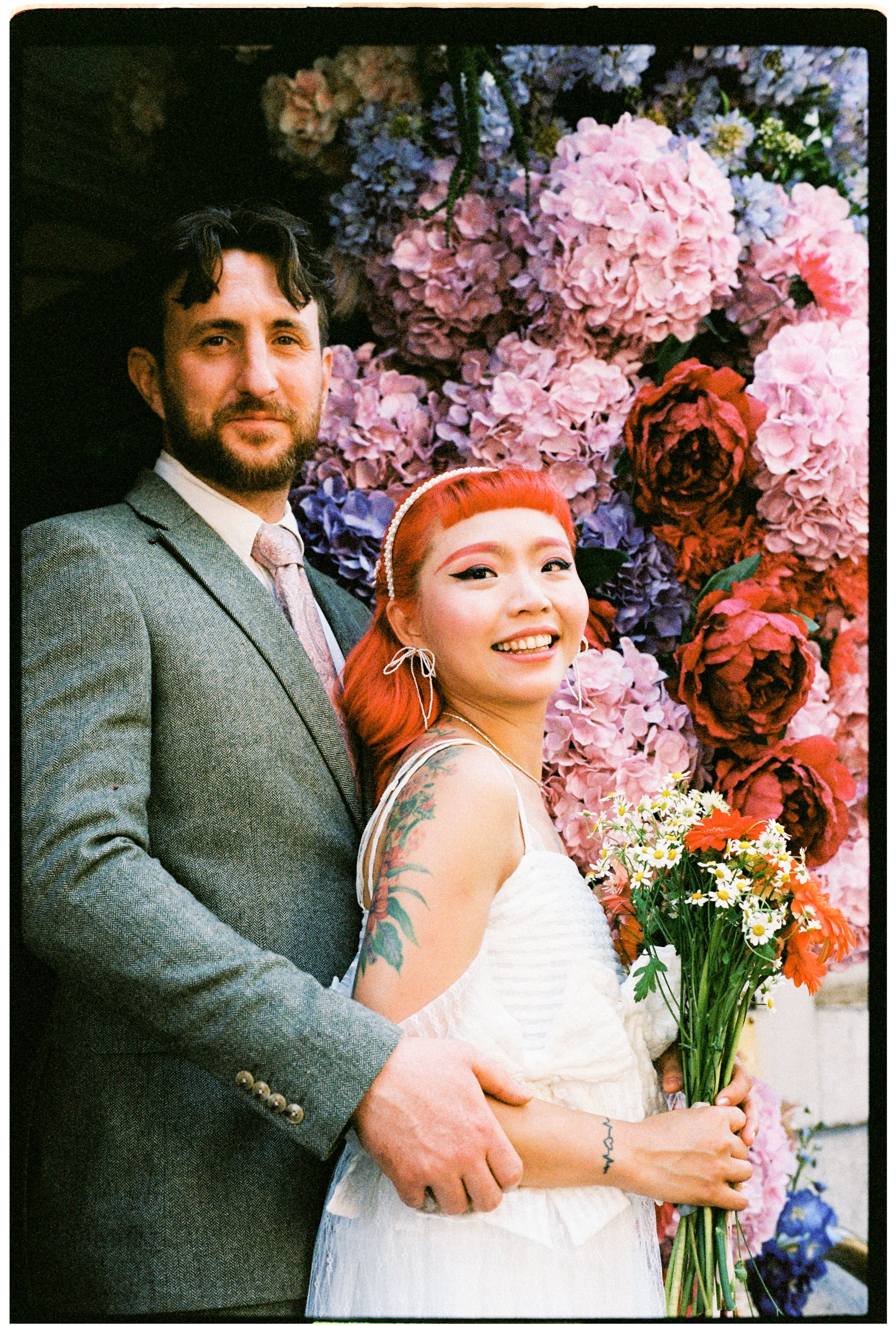A man with dark hair and beard wearing a suit, and a woman with bright red hair and tattoos on her arm holding a bouquet of orange and white flowers, standing in front of a large floral backdrop of pink and purple hydrangeas and roses.