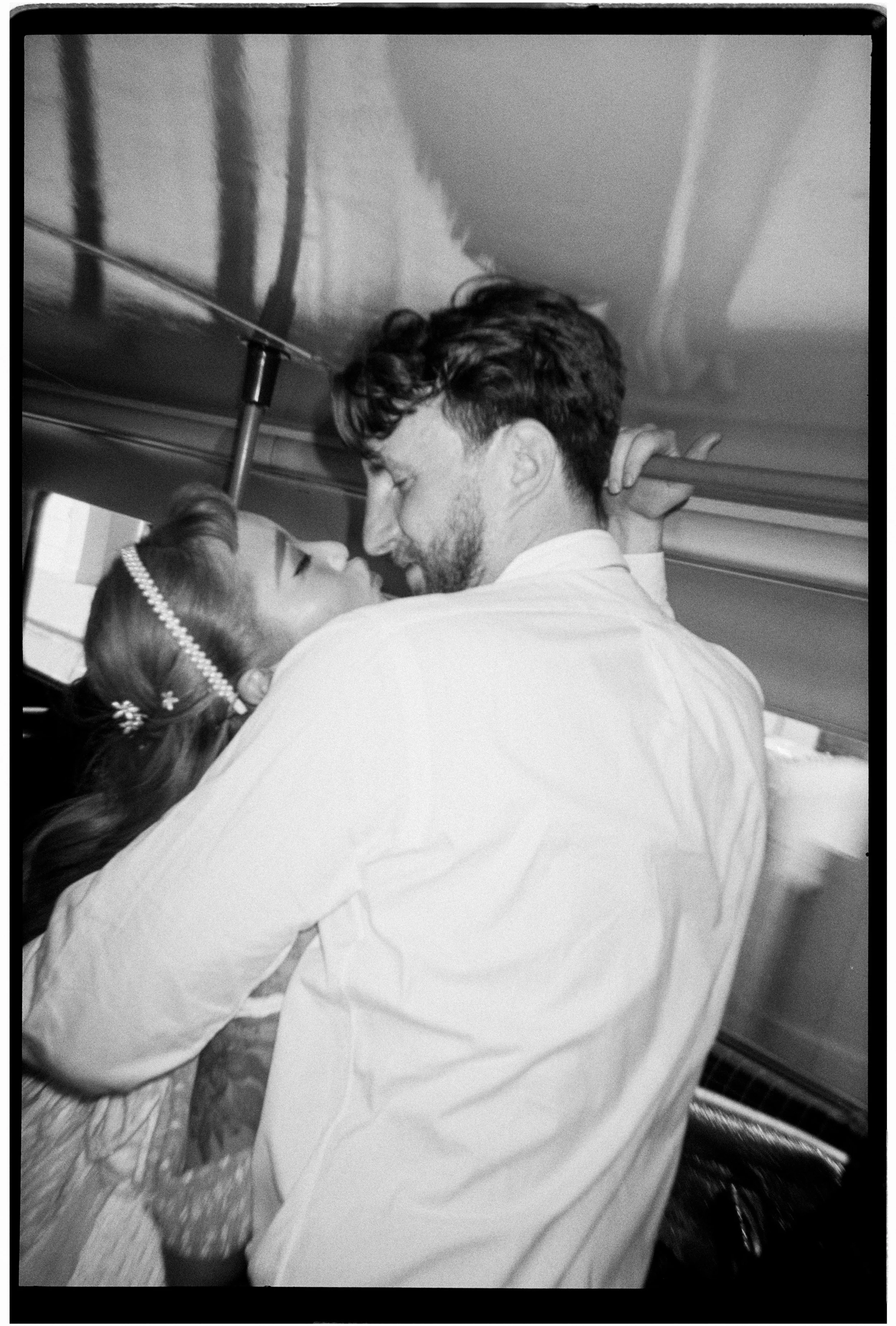 A couple sharing a romantic kiss inside a train, with the woman wearing a pearl headband and a floral sleeve dress, and the man with a beard and curly hair wearing a light-colored shirt.