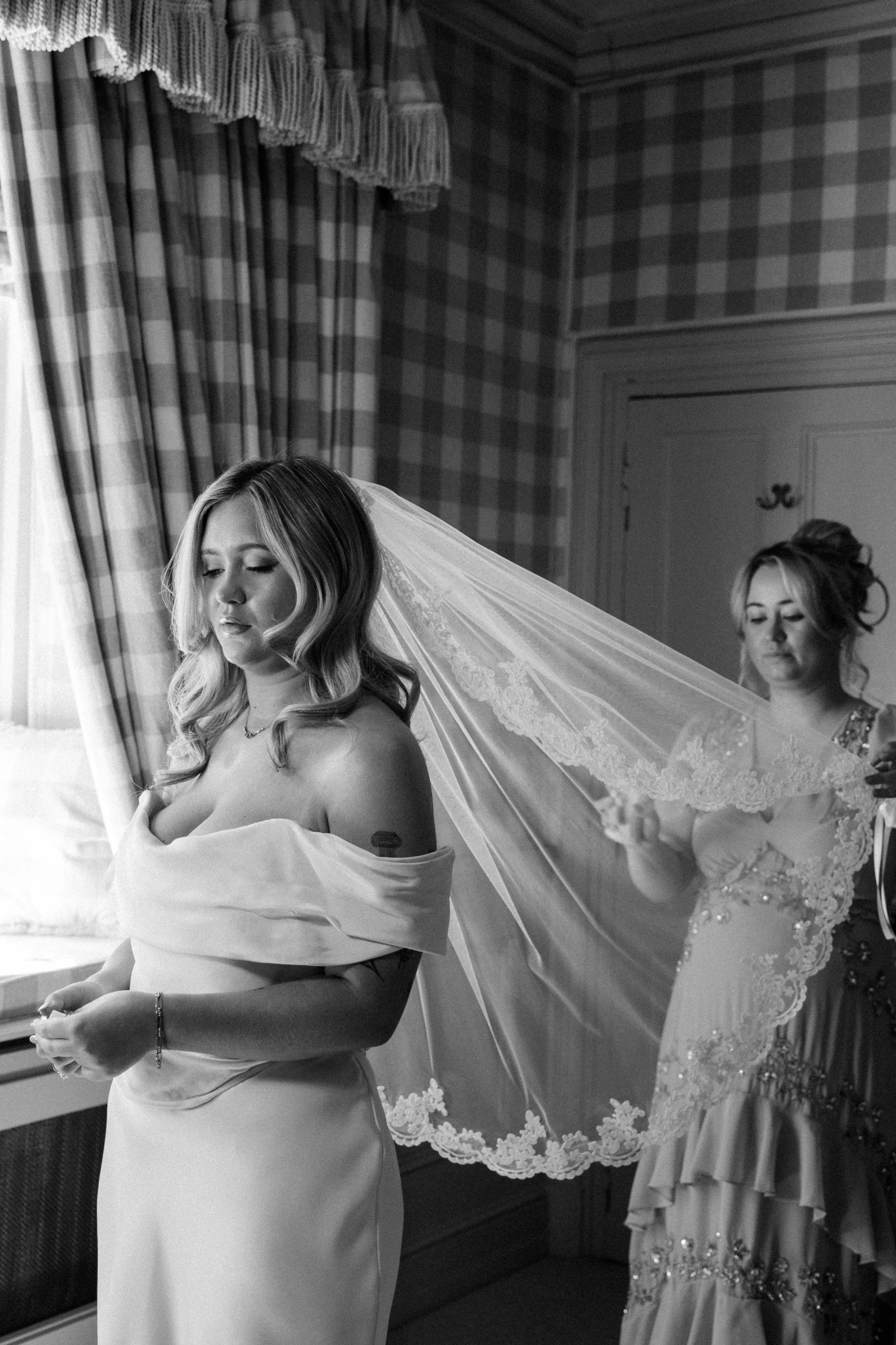 Two women, one with a wedding dress and veil and the other in an elegant gown, preparing for a wedding in a room with patterned curtains and decorated walls.