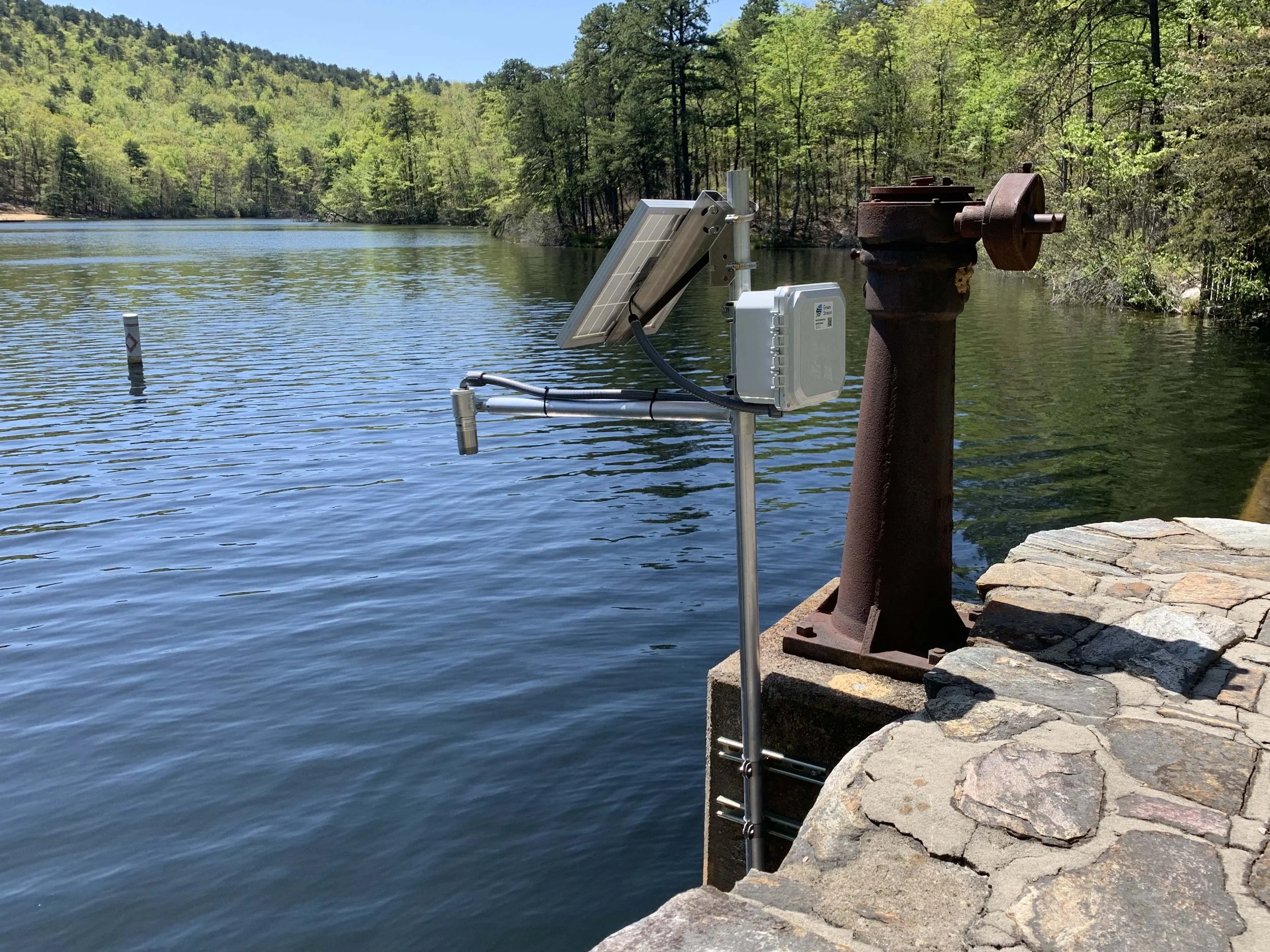 A water outlet pipe with a solar panel and a control box, situated on a stone and concrete embankment beside a river, surrounded by trees and hills under a clear blue sky.