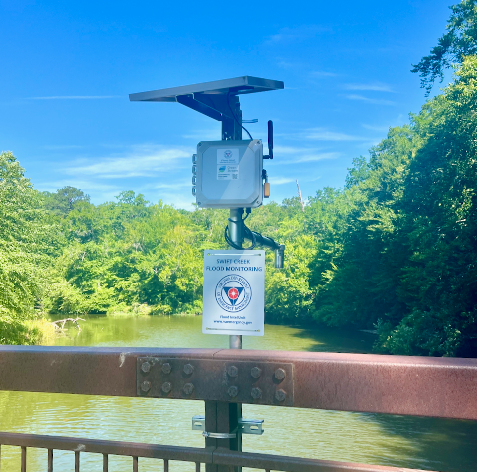 Flood monitoring device on a bridge over Swift Creek, surrounded by green trees on a sunny day.