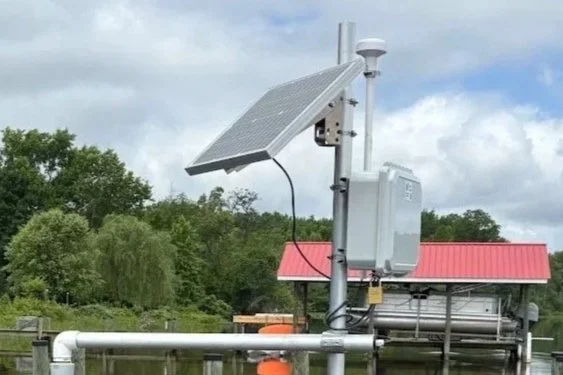A solar-powered weather station with a solar panel, an antenna, and electronic equipment installed on a metal pole near a body of water.