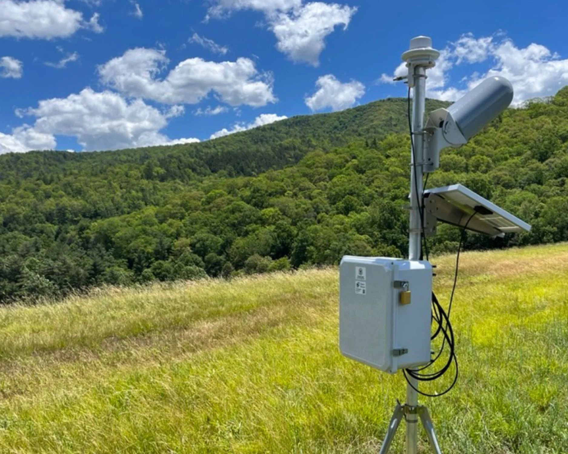 A precipitation monitoring station with sensors and equipment mounted on a pole in a grassy field, with a backdrop of green hills, trees, and a partly cloudy blue sky.