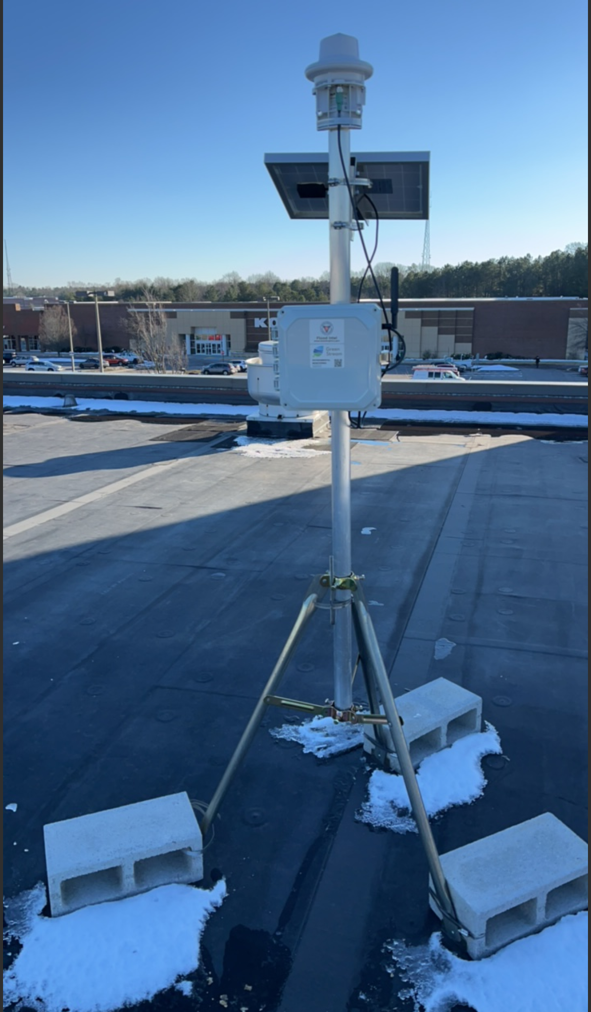 A weather station on a rooftop with solar panel, an anemometer, and other sensors, secured with cinder blocks and snow.
