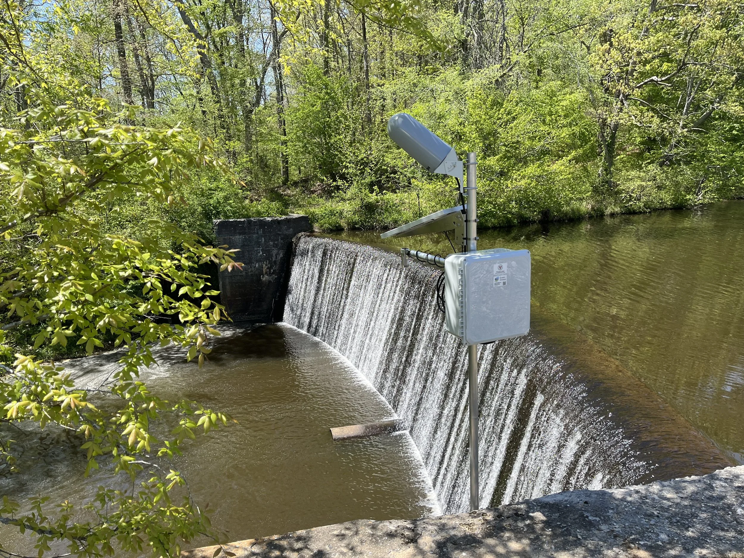 A water level sensor monitors the outflow of a high risk dam, set in a forested area with green trees and a stream.