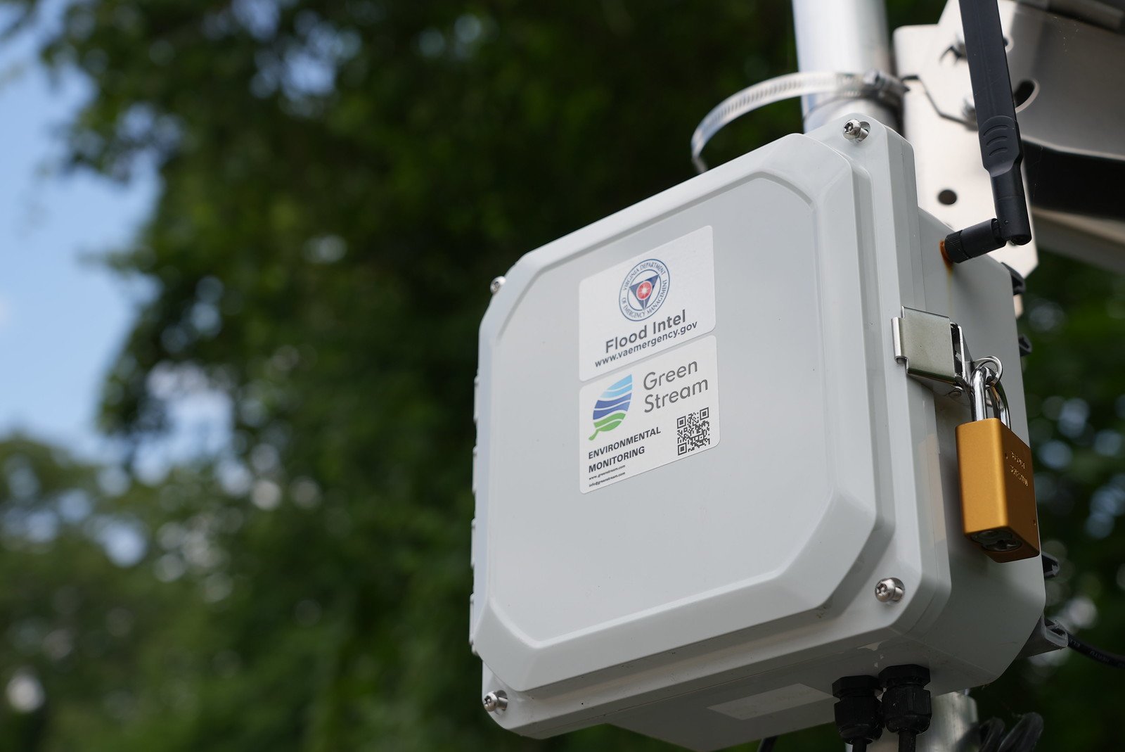 A Green Stream Corbo Environmental Transceiver mounted outdoors with green trees in the background. The device has labels for Flood Intel, Green Stream, and an environmental monitoring QR code.