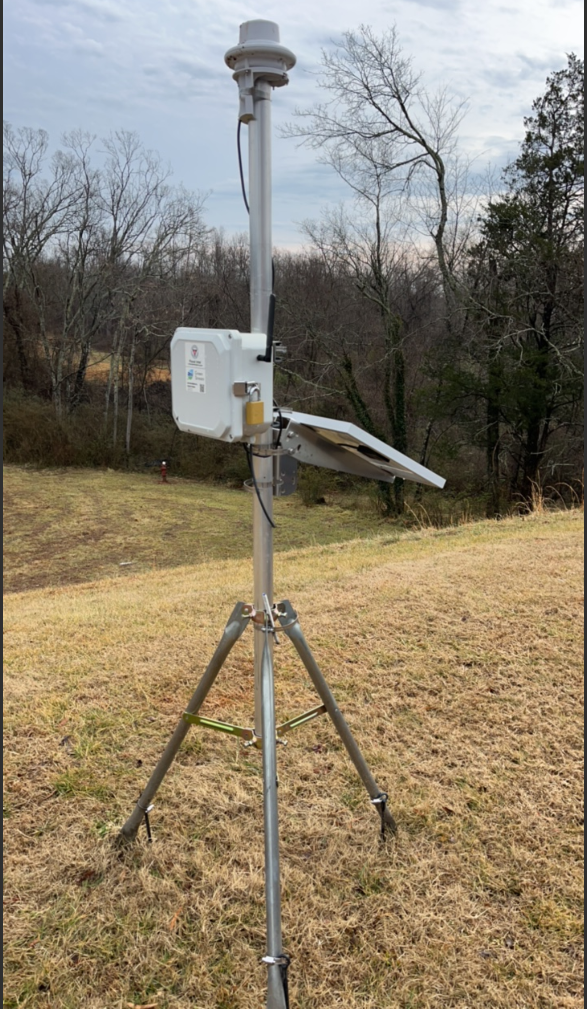 A precipitation monitoring station with sensors and antennas mounted on a tripod in an open field with trees and a cloudy sky background.