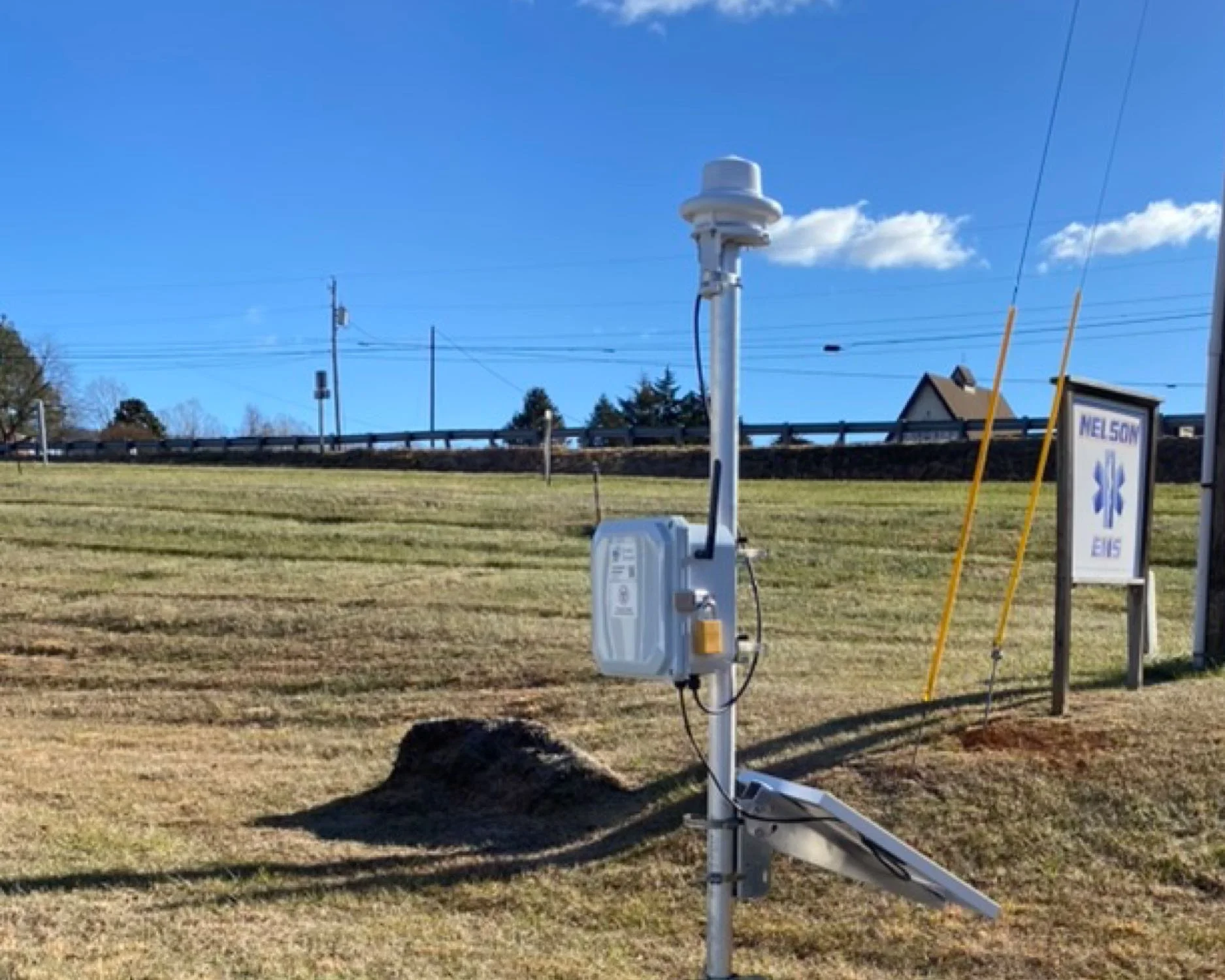A weather station with sensors and a sign that reads 'Nelson EMS' in an open grassy area under a blue sky with a few clouds.