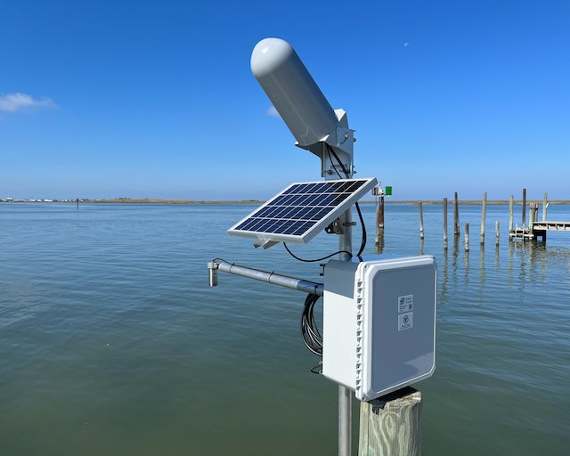 A weather station with a solar panel, an anemometer, and a control box, located near the water on a dock.