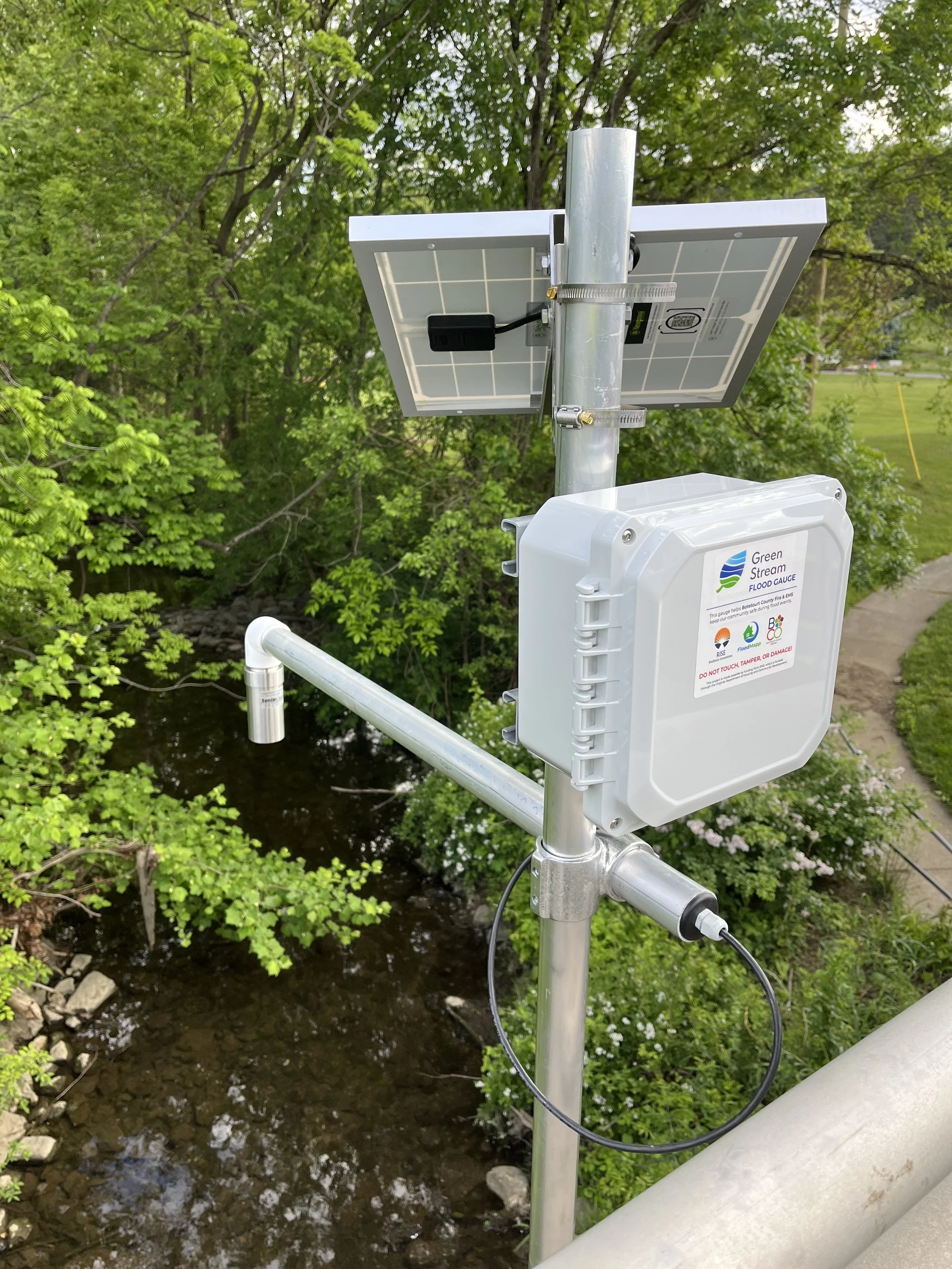 A solar-powered flood gauge installed with a white rectangular box, a solar panel, and a sensor cable over a creek with green trees in the background.