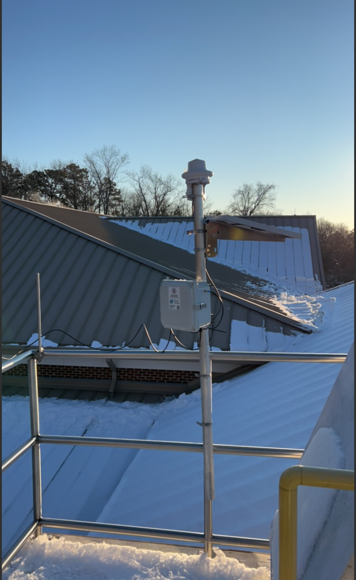 Installation of a precipitation monitoring station on a rooftop with snow, metal railings, a chimney, and a sunny sky.