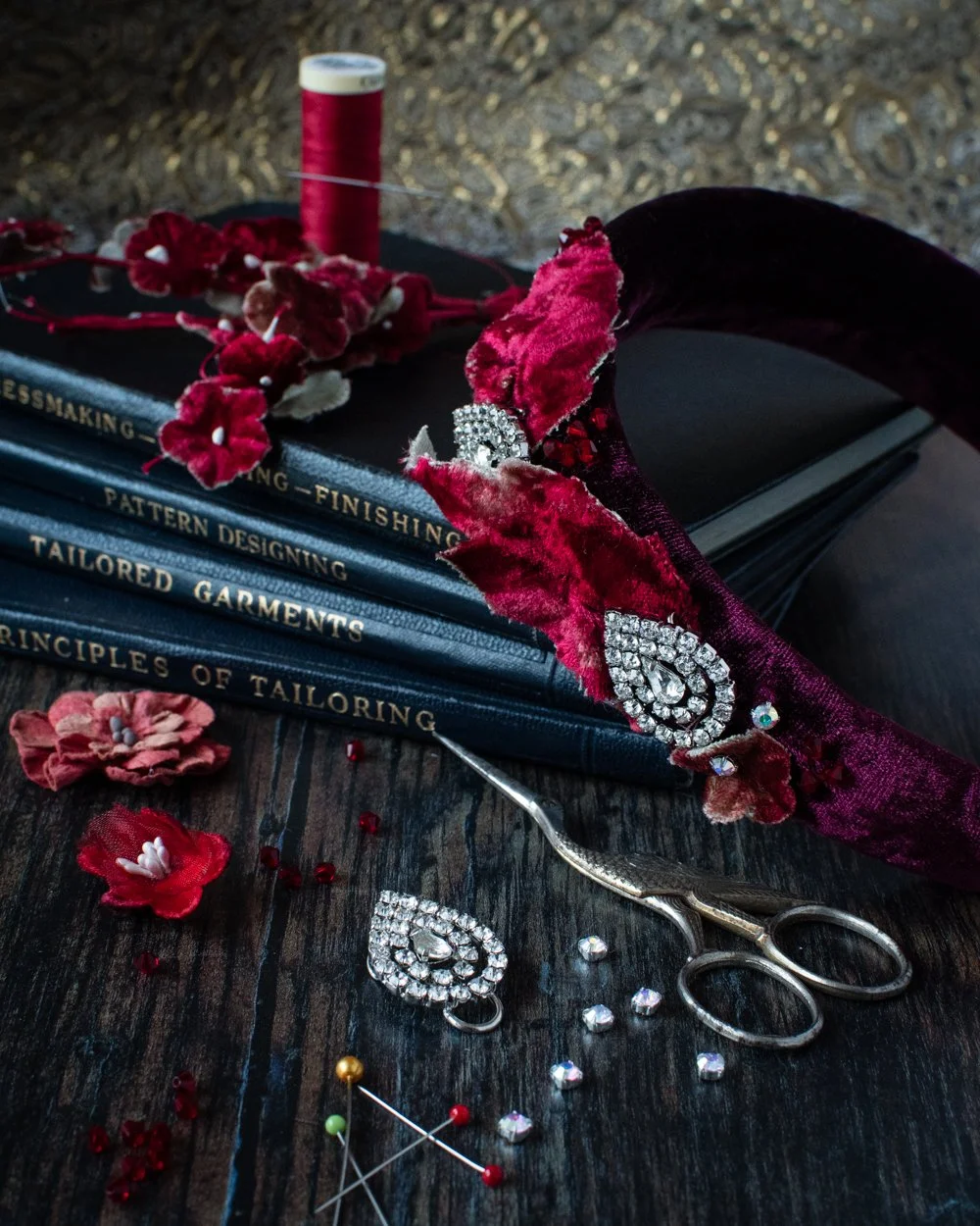 An hand sewn embellished velvet headband with velvet leaves and diamante brooches on a pile of vintage dressmaking books and a reel of red thread with a sewing needle