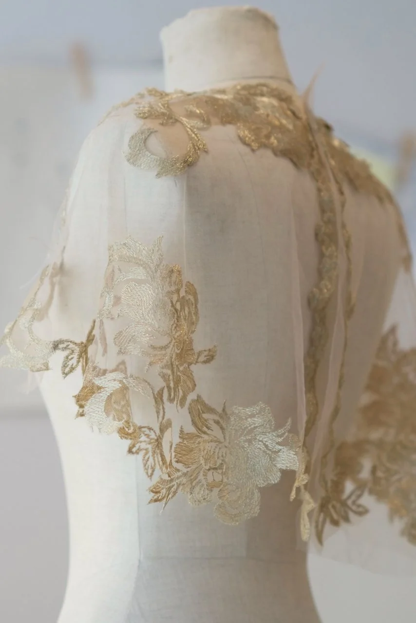 A photograph of a mannequin on a sewing table in the atelier, with hand-sewn lace appliqué on nude tulle.
