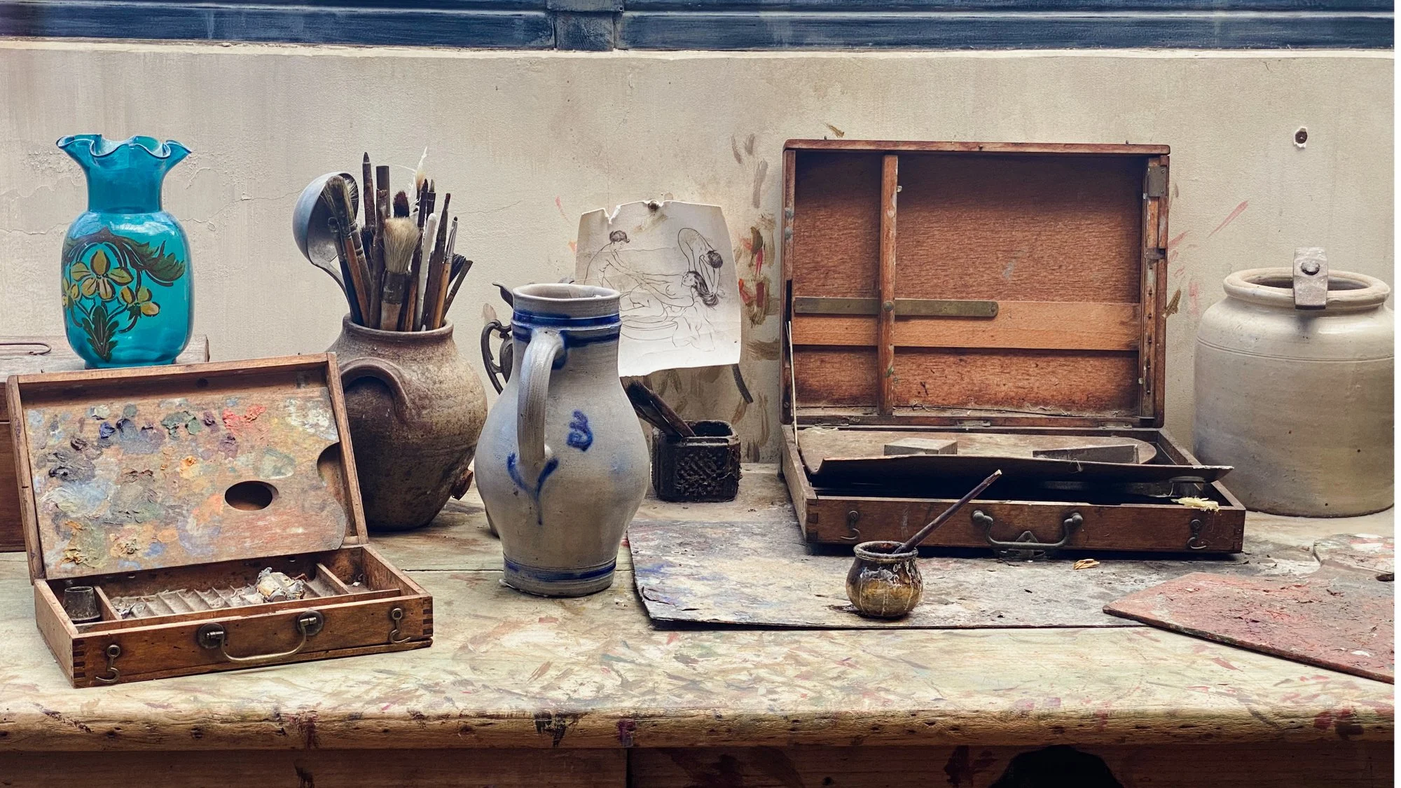 Oil paint palettes and a jar full of artists paintbrushes on an old wooden table in Suzanne Valadon's studio.