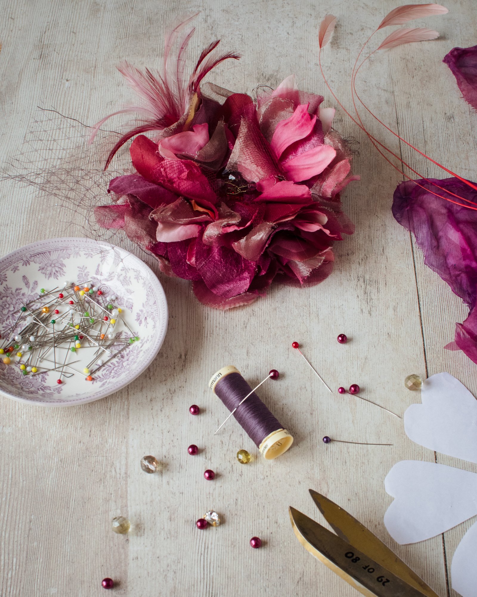 A craft workspace featuring a decorative flower made of pink and red fabric, various sewing pins with colorful heads, a spool of purple thread, a pair of gold scissors, and a paper pattern on a light wooden surface.
