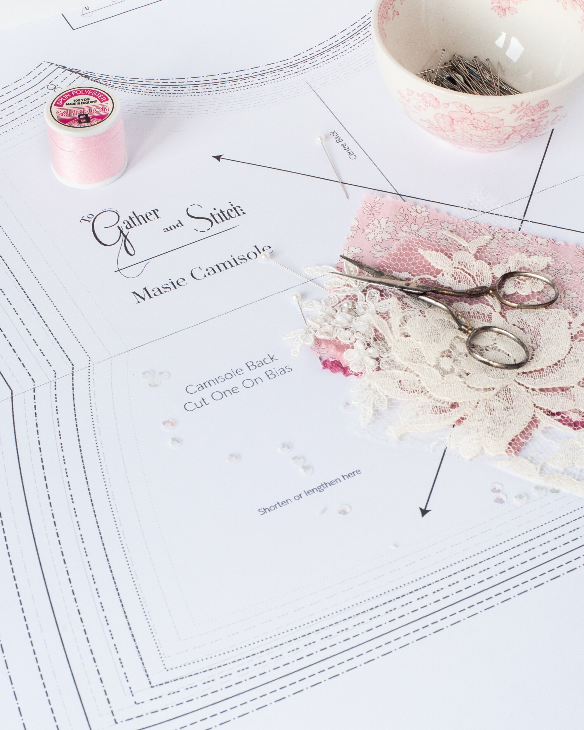 A sewing pattern, sewing equipment including a needle, scissors, pink lace, a spool of pink thread, and a bowl of sewing pins on top of the pattern.