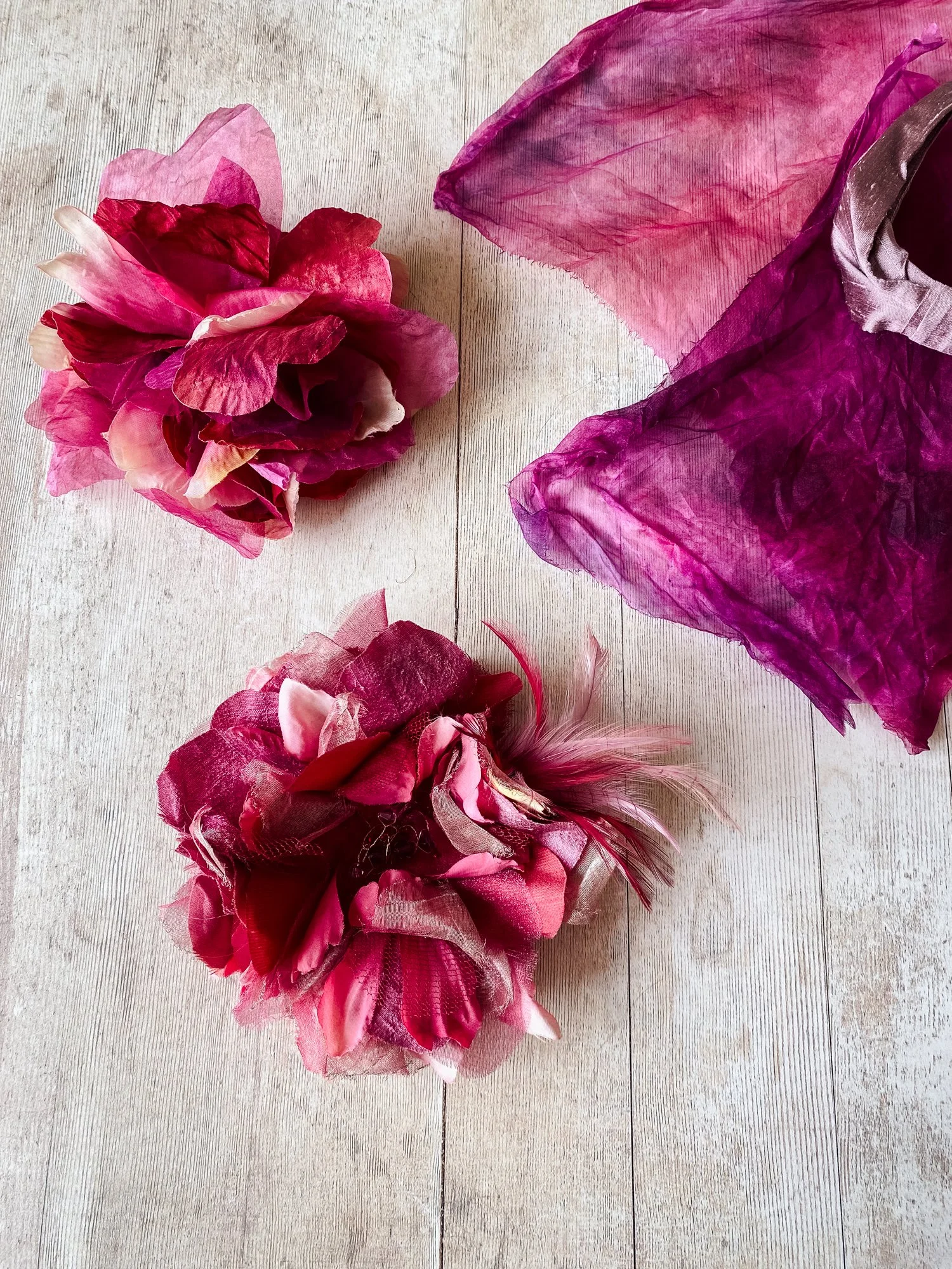 Three pink and red fabric flower corsages with feathers placed on a light wood surface, with a large pink and purple tulle bow on the top right.