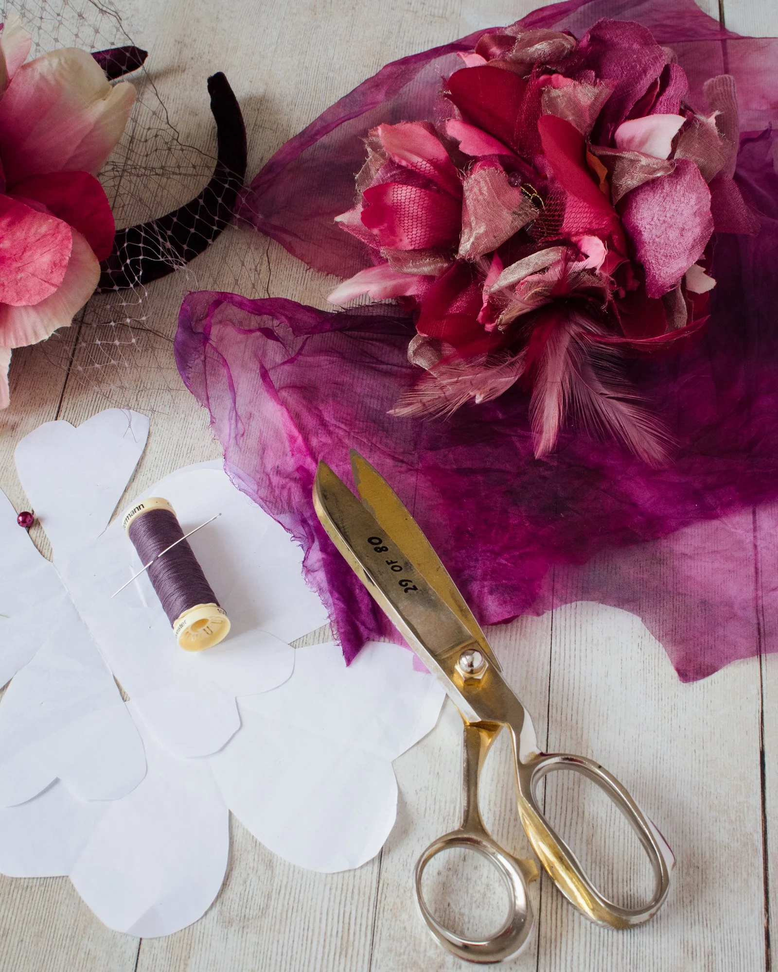 Wedding bouquet with pink and red flowers wrapped in pink and gold mesh fabric, placed on a wooden surface. There are also sewing tools including a pair of golden scissors, a spool of purple thread, and cutout paper hearts near the bouquet.