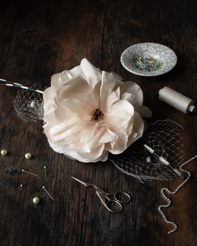 A large white fabric flower on a dark wooden surface, a pair of gold scissors, pins with white and pearl-colored heads, a spool of white thread, a pearl necklace, and small decorative mesh pieces.