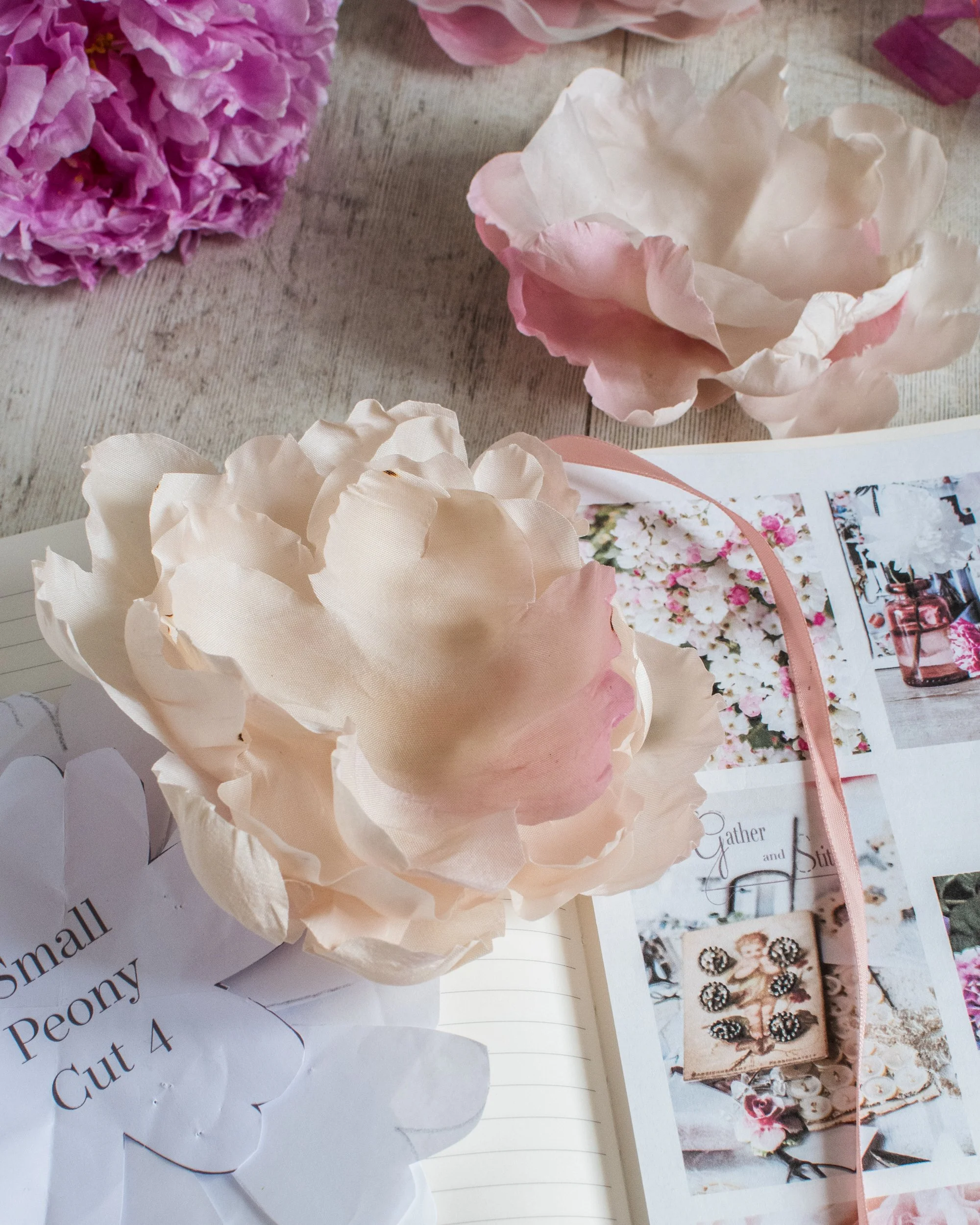 Pink and white artificial flowers arranged on a wooden surface with a photo album or scrapbook and printed paper with floral images and text.
