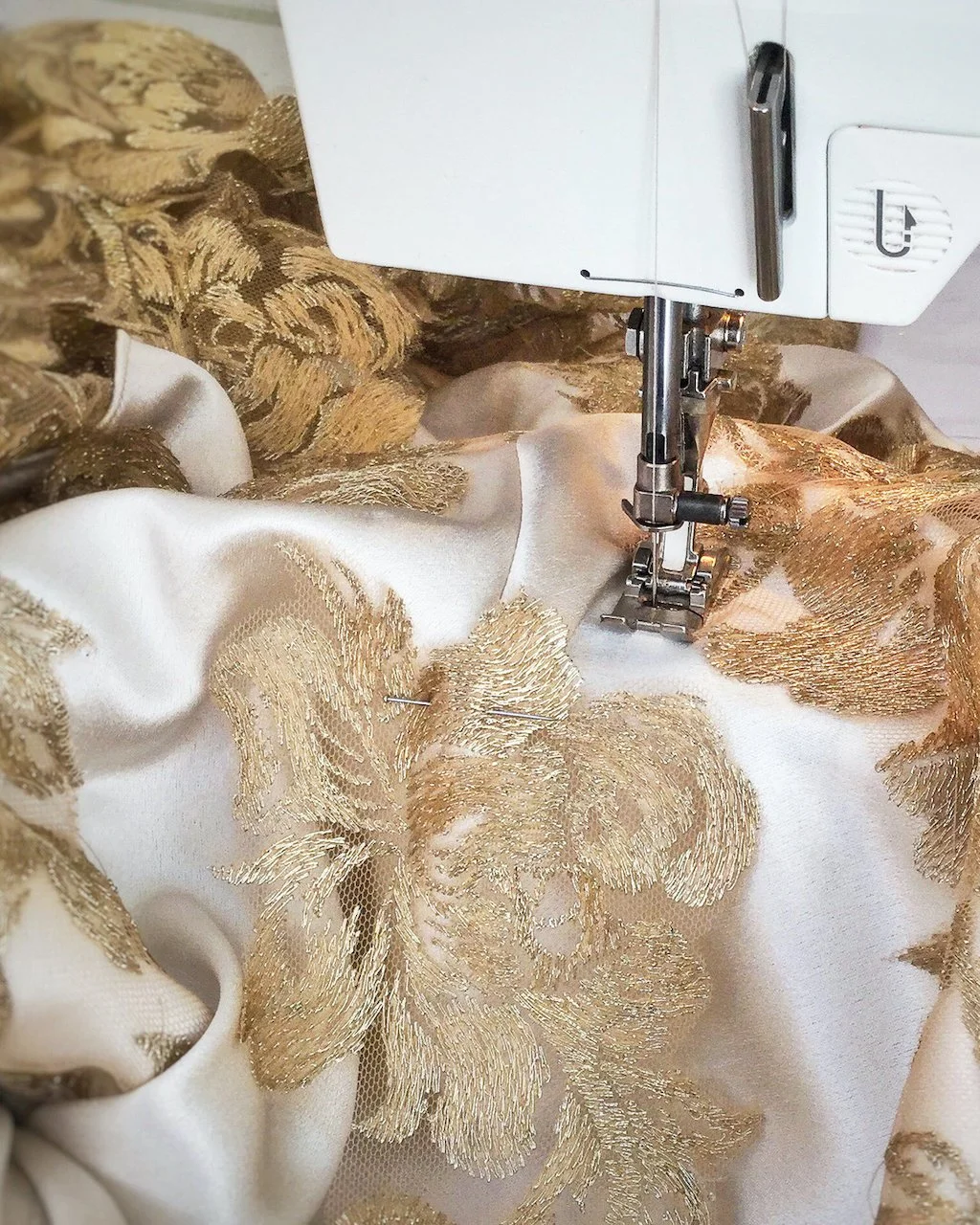 Photograph of using a sewing machine to appliqué flower motifs cut from gold lace fabric to blush silk satin.