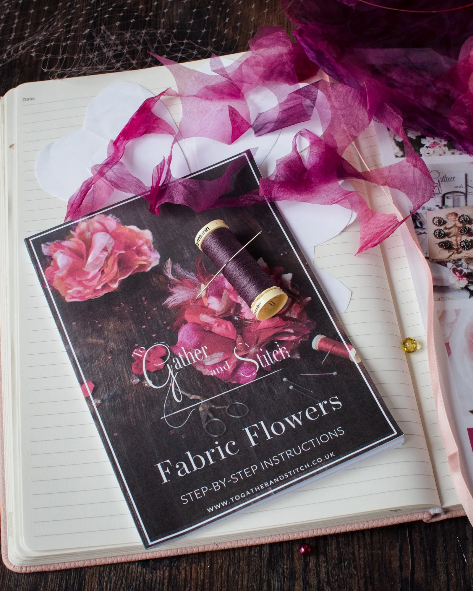 A collection of craft materials on an open notebook, including a flyer titled "Gather and Stitch," pink and purple tissue paper, a spool of purple thread, a sewing needle, and a few decorative gems on a wooden surface.