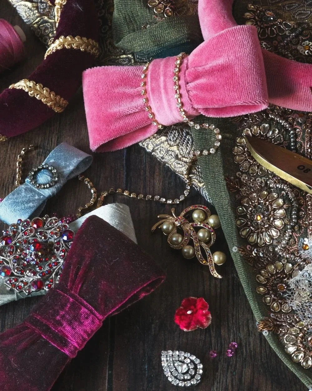 A dark wooden tabletop in the sewing studio, with couture bows, fabric flowers, beaded trimmings and vintage brooches scattered upon it.