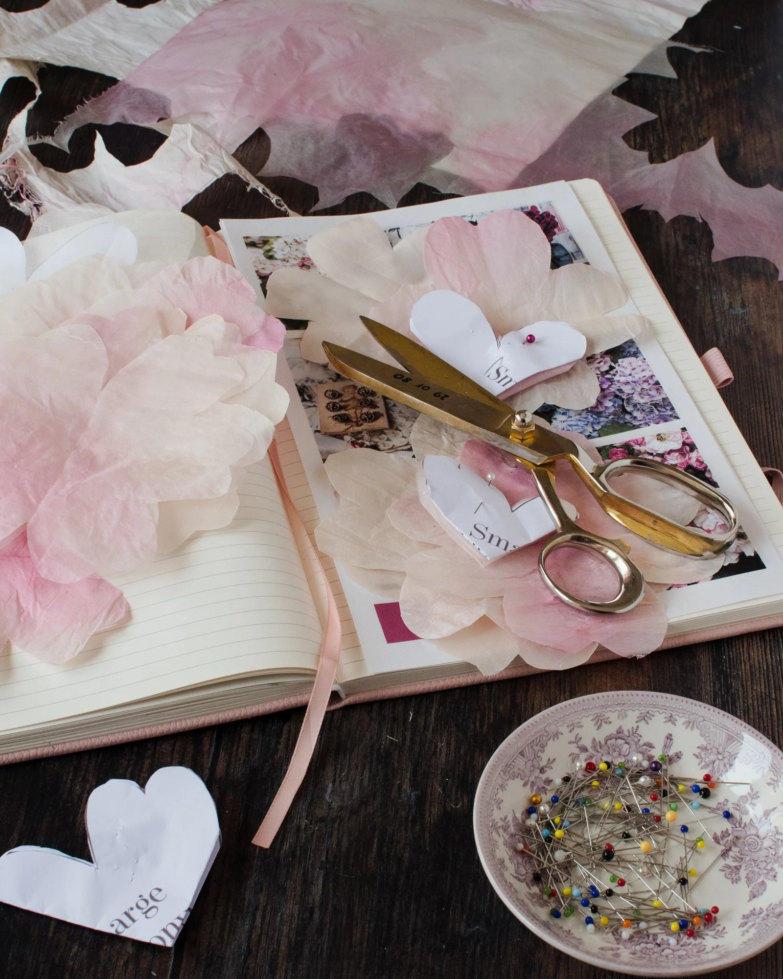 A craft workspace with pink and white tissue paper flowers, scattered paper hearts, a pair of gold-handled scissors, a scrapbook or planner with photos, and a decorative plate filled with colorful sewing pins on a wooden surface.