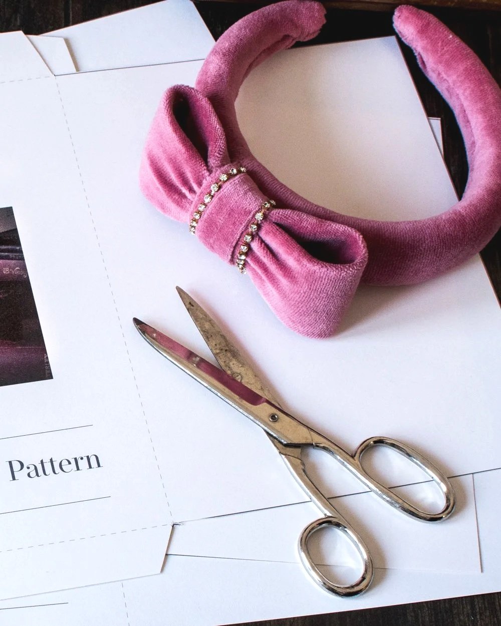 Pink velvet headband with a bow and rhinestones, silver scissors, and pattern papers on a dark surface.