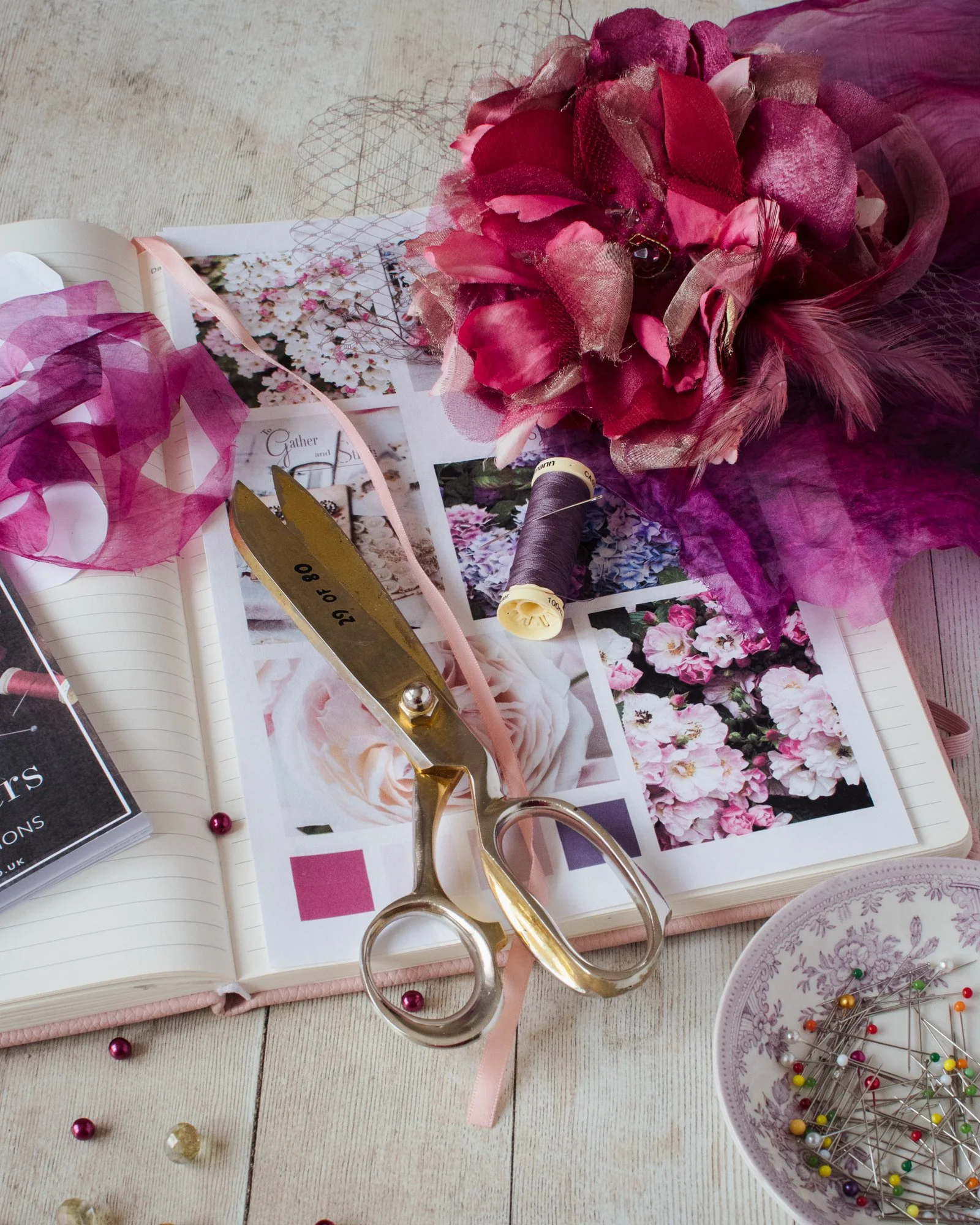 A scrapbook with pink flowers, sewing scissors, thread, and colorful pins on a wooden surface.