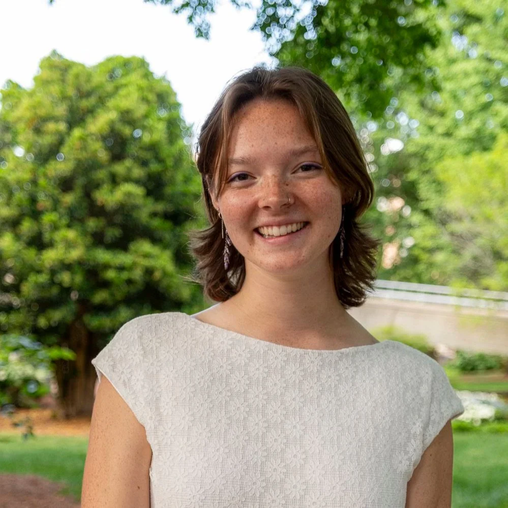 A young woman with shoulder-length brown hair, freckles, and earrings, smiling outdoors with trees and greenery in the background.