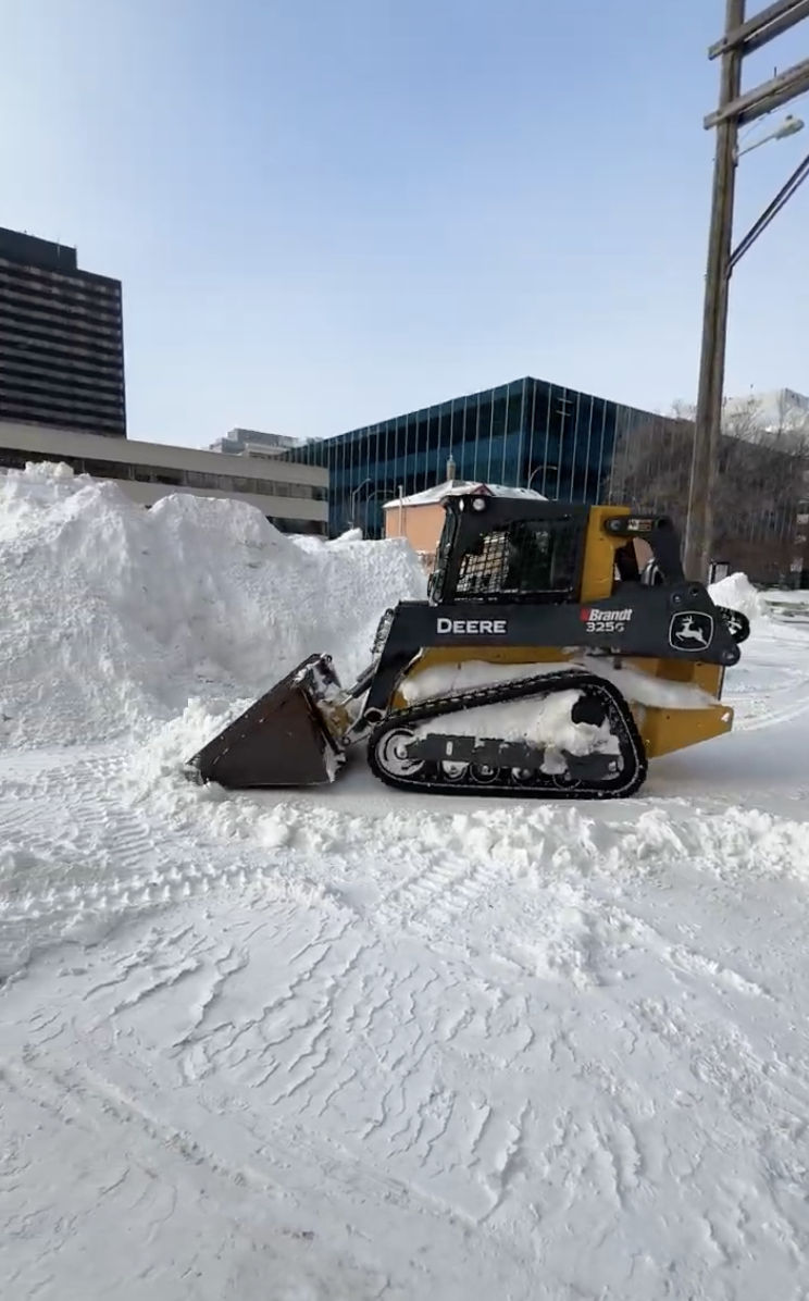 A John Deere snow blower clearing snow on a city street with buildings and power lines in the background.