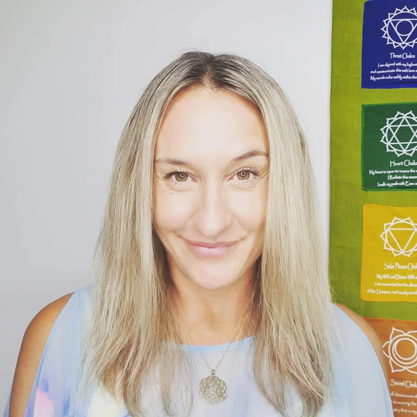 A woman with long blonde hair smiling at the camera, wearing a light blue top and a gold pendant necklace, with colorful chakra flags on the wall behind her.