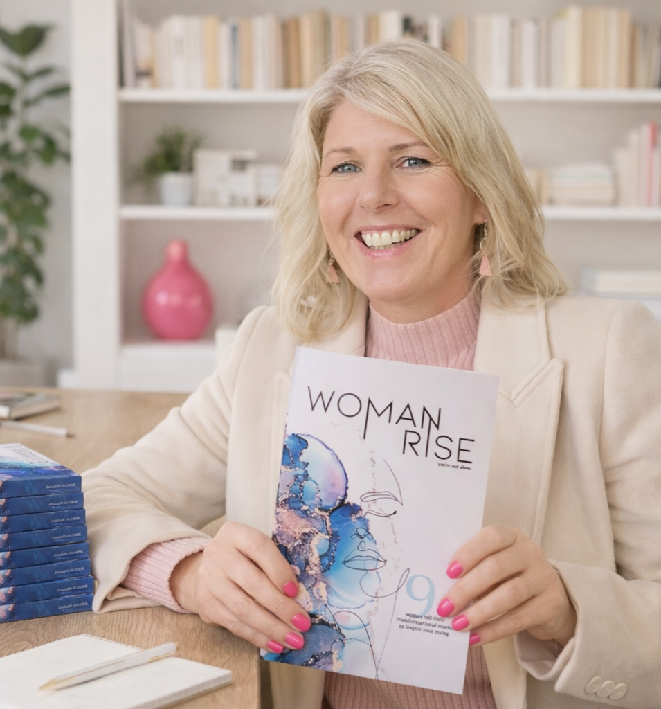 Founder of Hot Mess Goddess, author mentor and writer  Sally Friday sitting at a table holding a book titled "Woman Rise" in a bright, well-decorated room with a bookshelf, a pink vase, and some plants in the background.