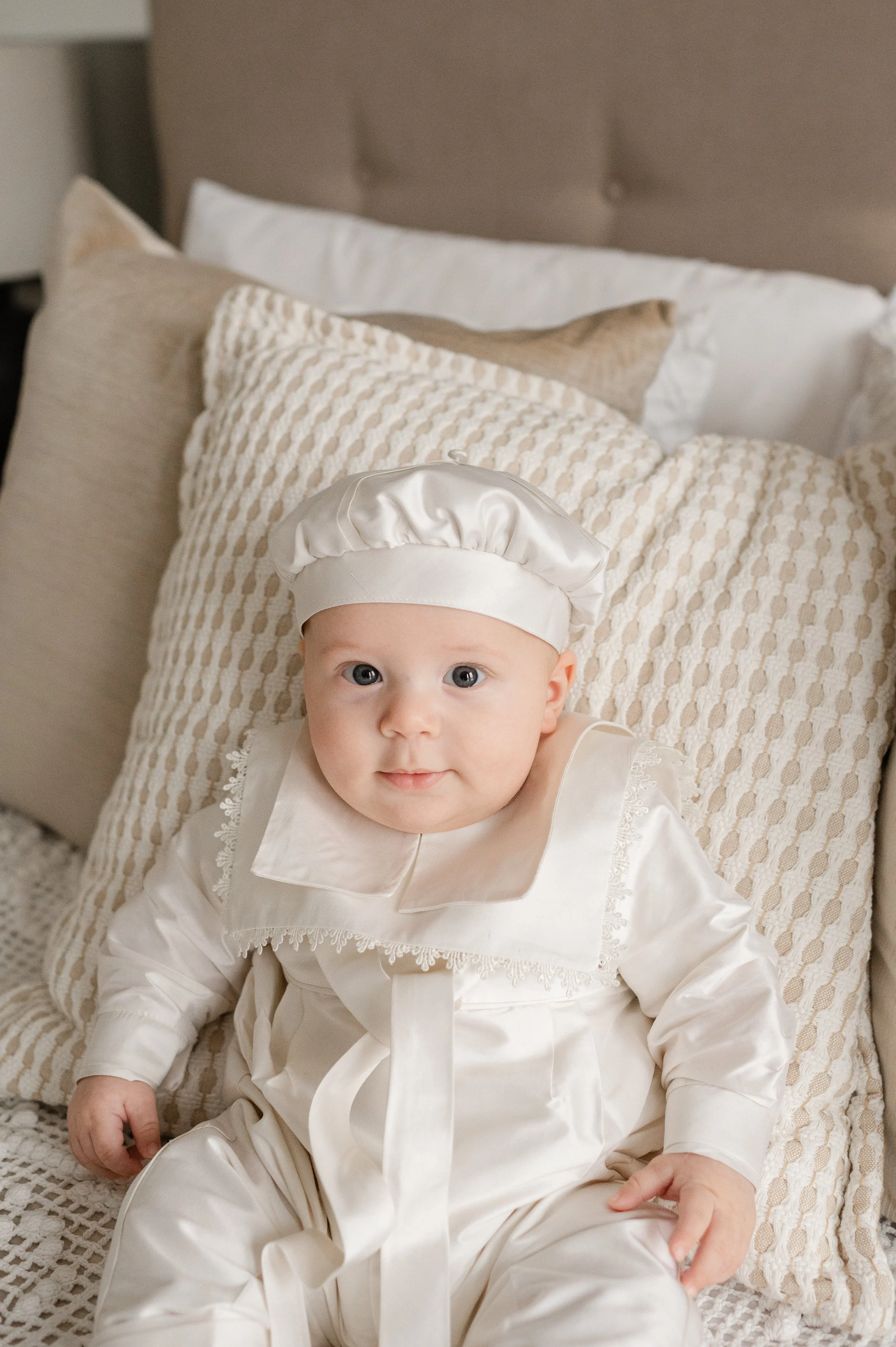 A baby sitting on a bed, dressed in a baptism outfit with a matching hat, surrounded by beige and white pillows. Toronto baptism photography