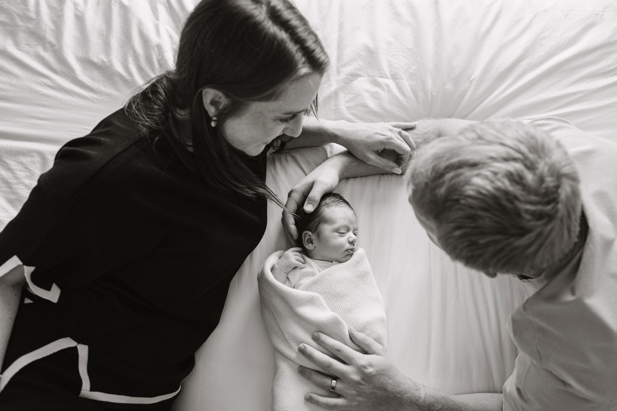 a mother and father looking at their newborn son, who is swaddled in a blanket. They are all laying on a bed together with the baby in the middle, black and white image. Newborn photography, newborn photographer, GTA newborn photography