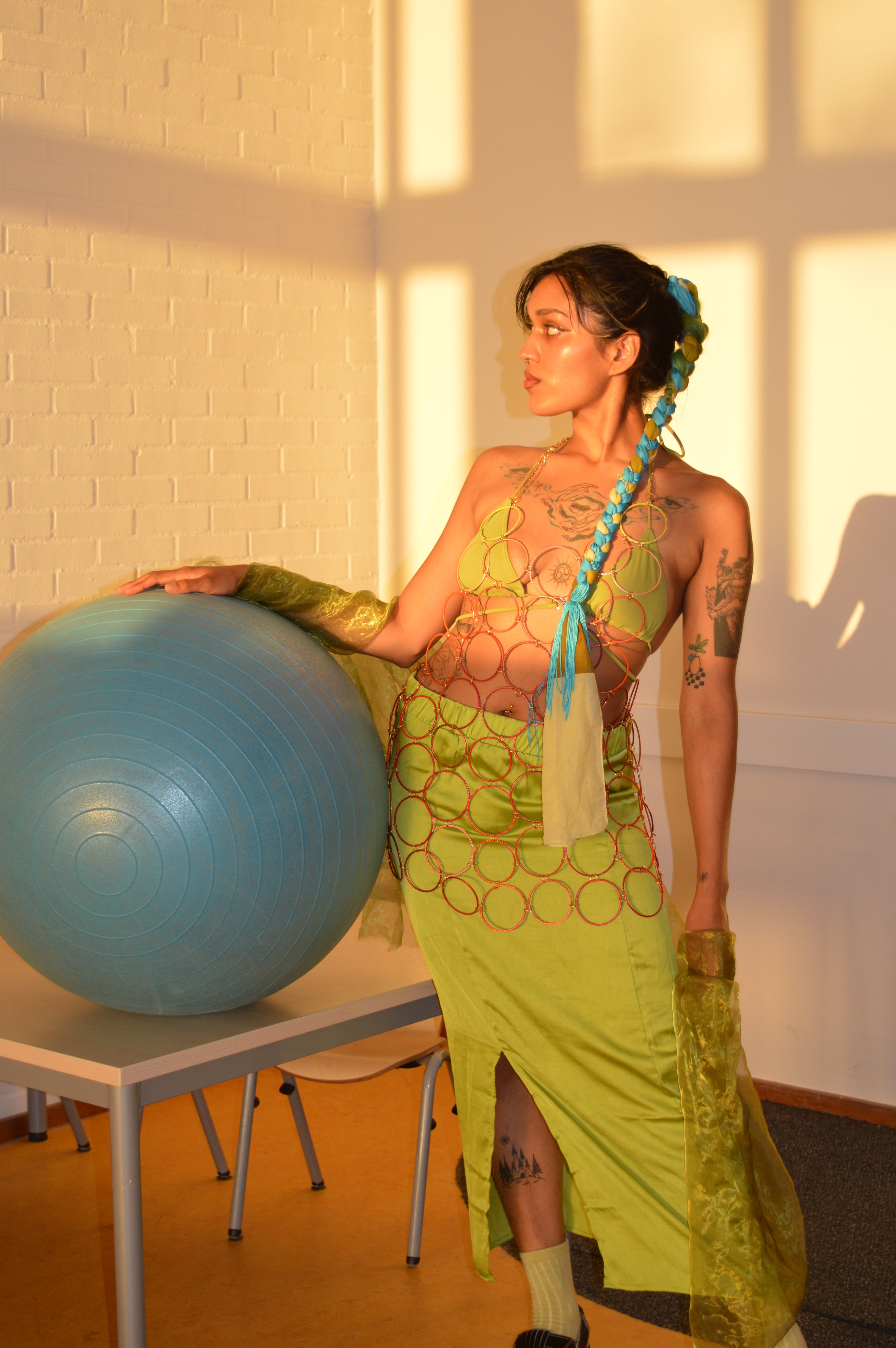 A woman stands next to a large blue exercise ball in a room with sunlight streaming through window shadows. She is wearing a fashionable outfit made of colorful rings, a yellow skirt, and matching accessories, with tattoos visible on her arms and leg