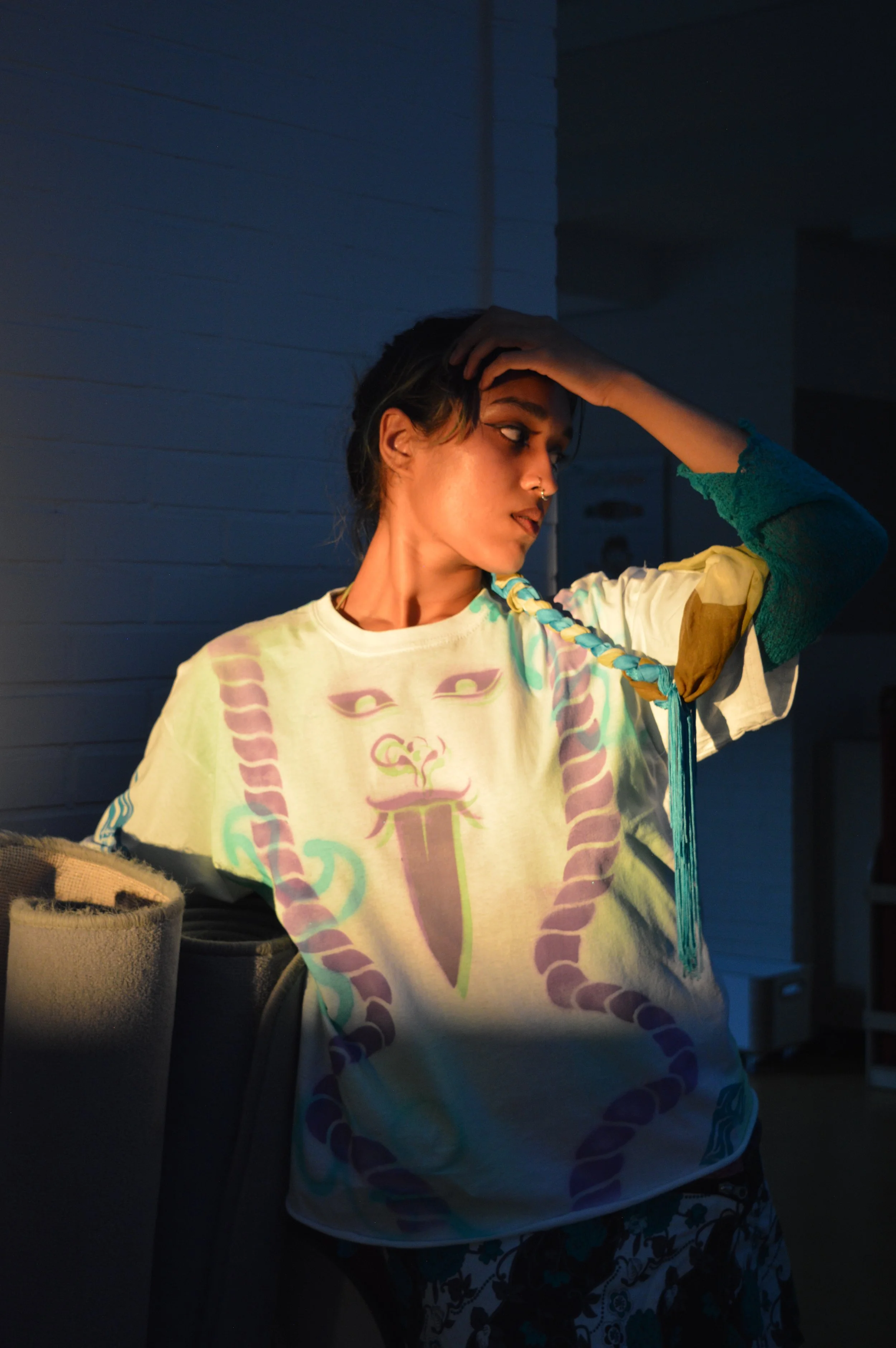 A young woman with dark hair and makeup, wearing a white shirt with a graphic design, stands in a dimly lit room with blue-tinted lighting. She is looking to her right, with her left hand on her head, and a braid with colored ribbons over her shoulde