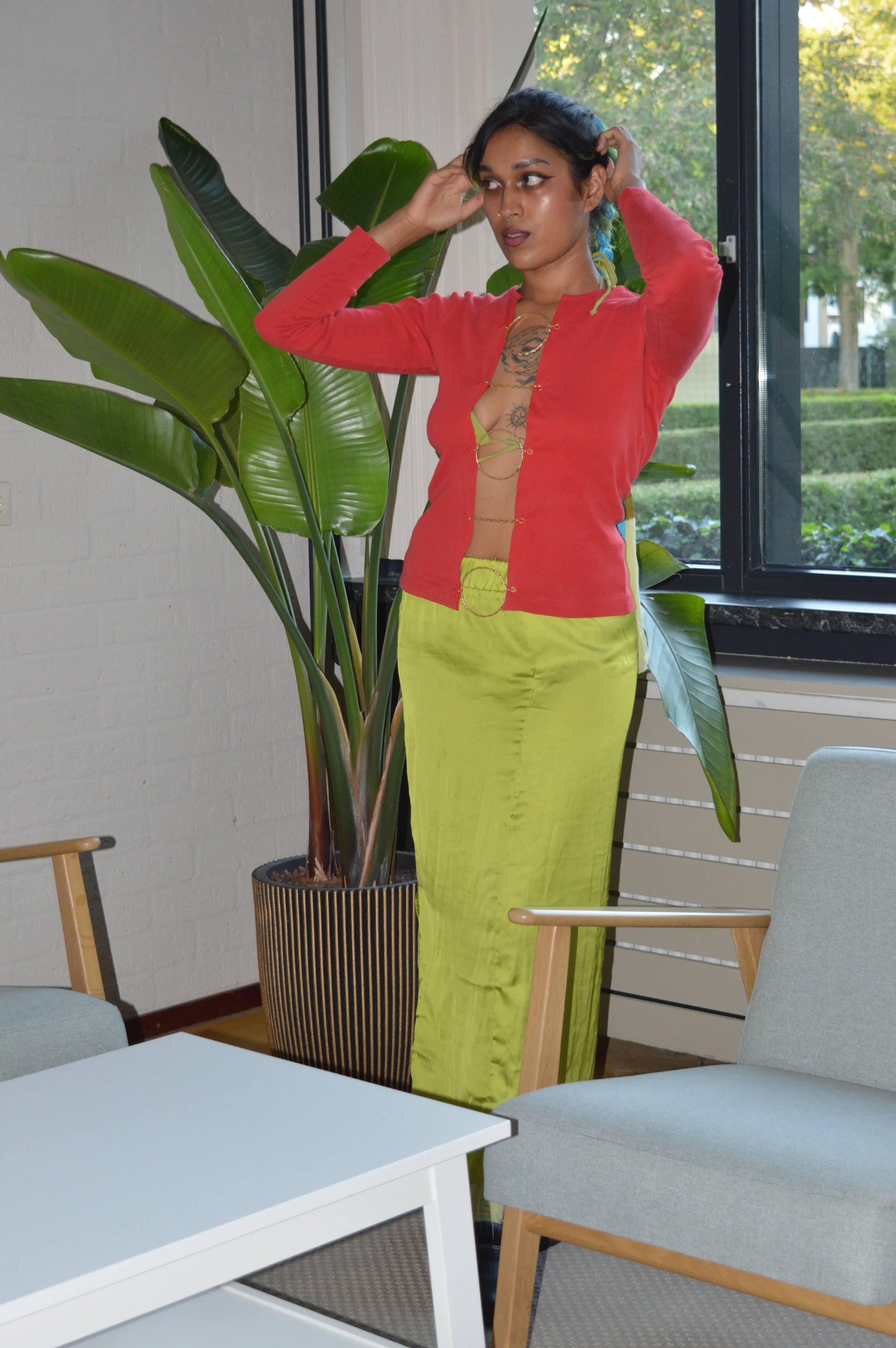 A woman with dark hair and tattoos stands indoors near a large potted plant at a table, wearing a colorful outfit, and adjusting her hair.