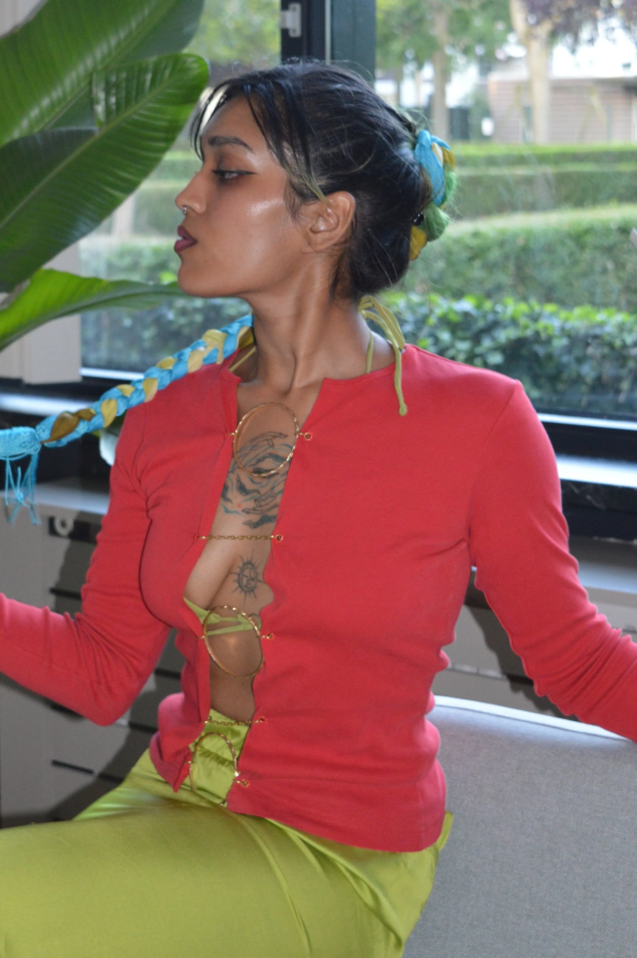 A woman with dark hair styled in a long, colorful braided ponytail, dressed in a red top with cutouts held together by gold rings, and bright yellow pants, sitting indoors near a large window with greenery outside.