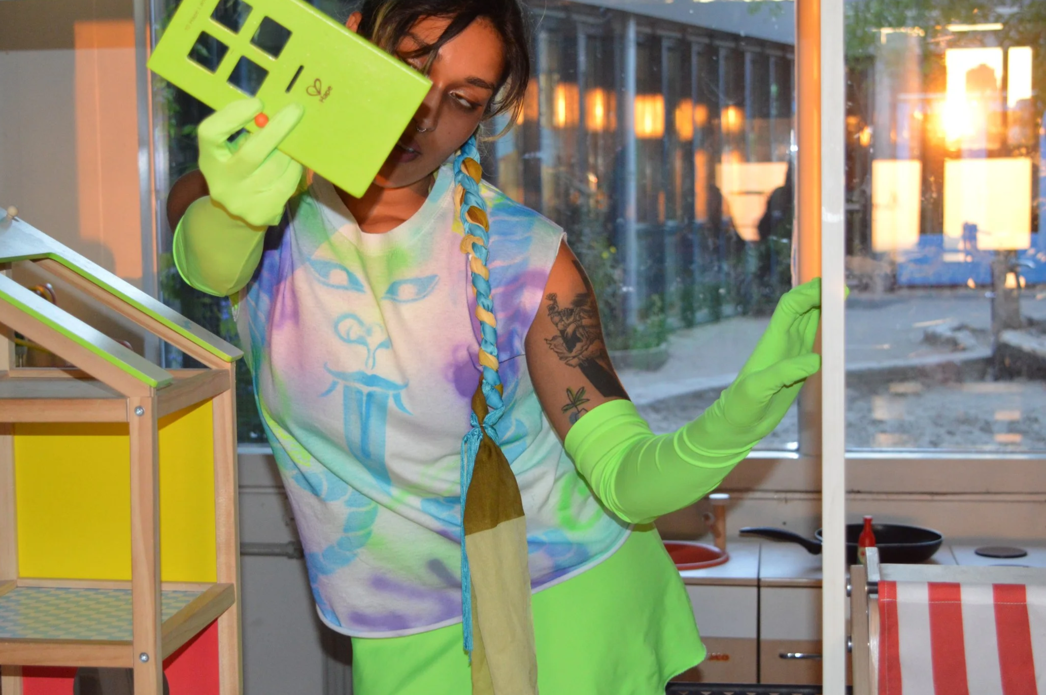 A woman with colorful hair braid, wearing green gloves and an airbrushed shirt, holding a yellow object with black cutouts and standing indoors next to a window and colorful shelves.