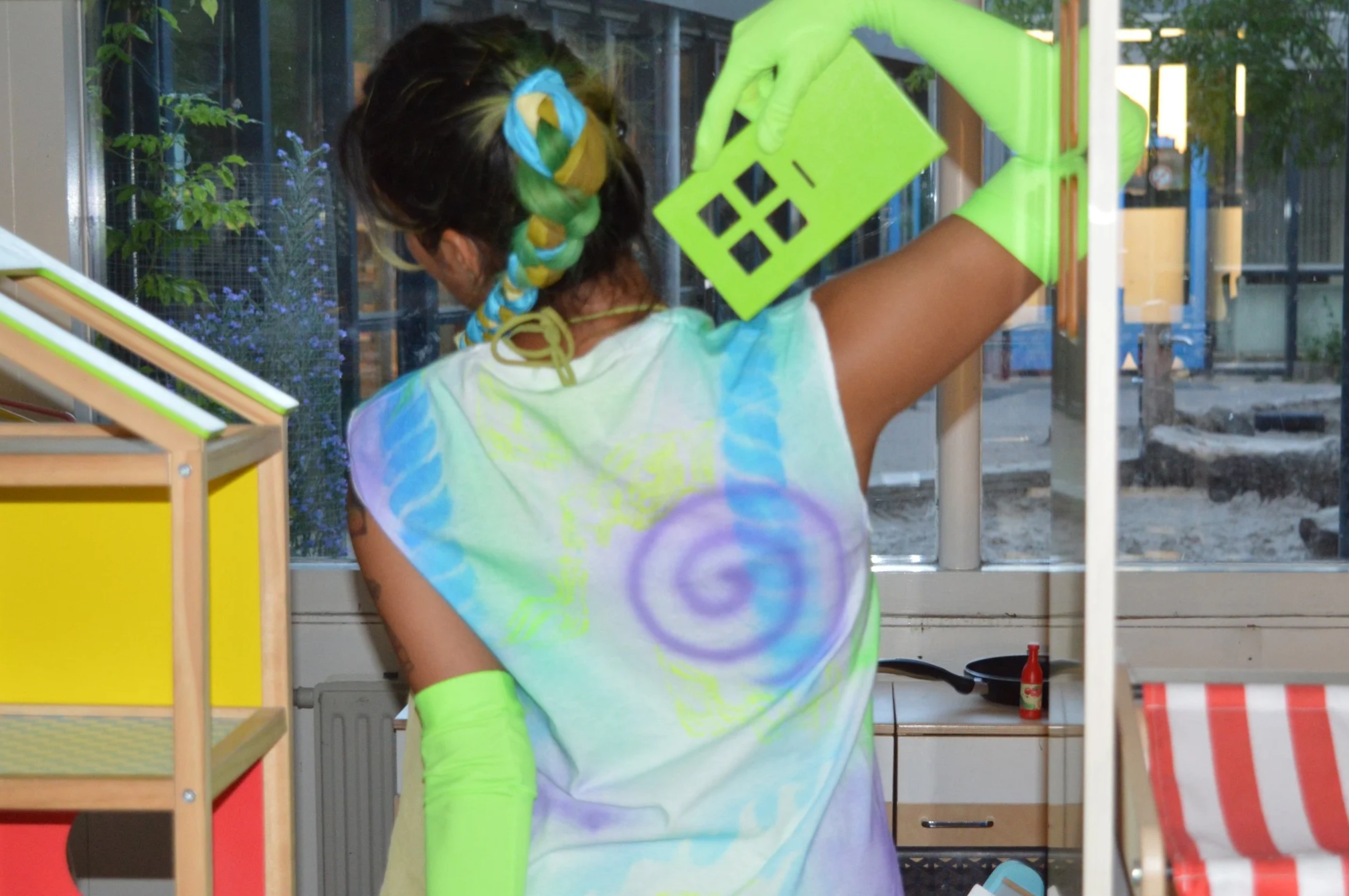 A person with multicolored braided hair, wearing bright green gloves and a colorful airbrushed sleeveless shirt with a spiral design, is standing inside a room near a window, holding a green plastic object.