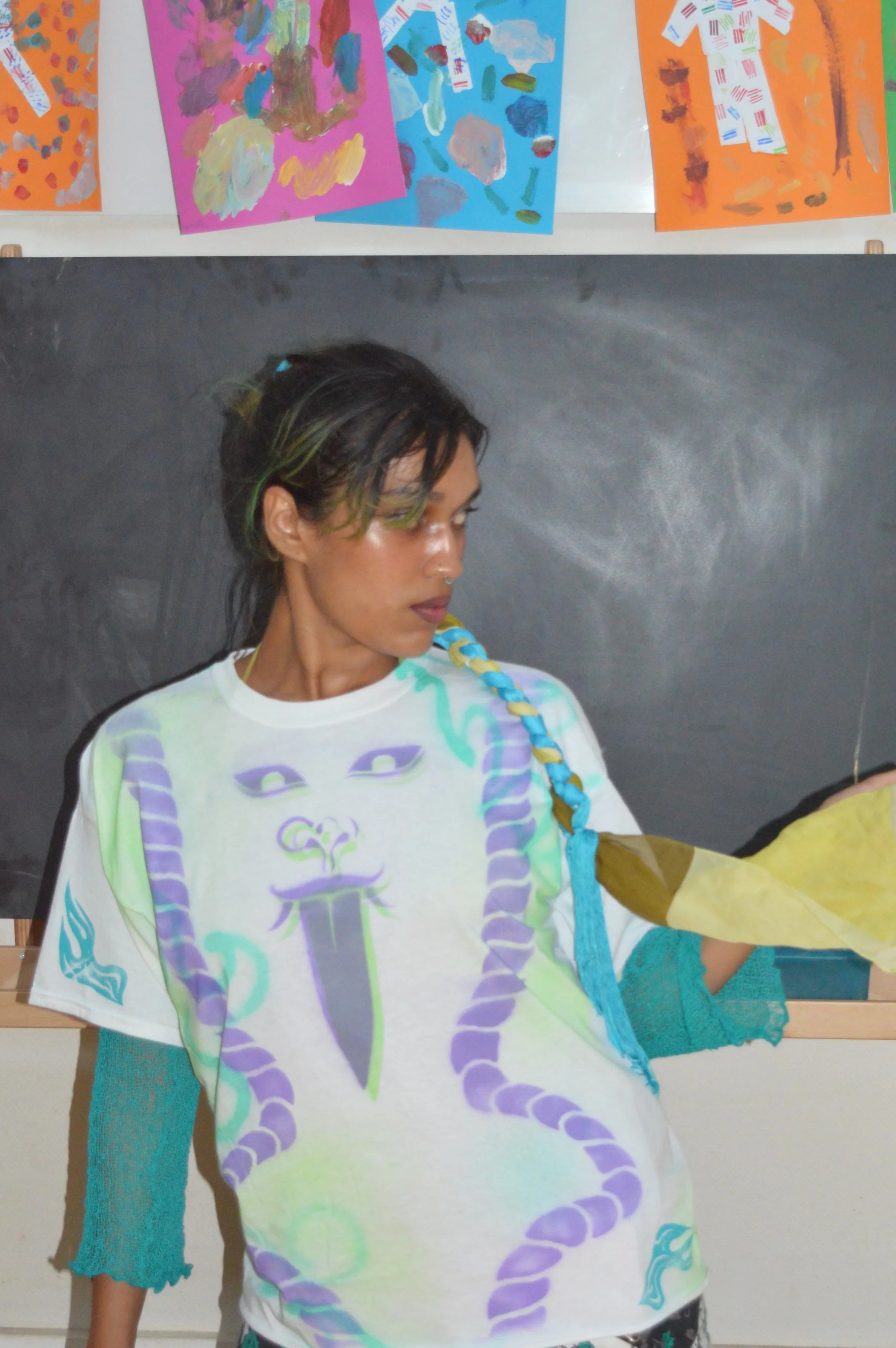 A young woman with dark hair and green highlights, wearing a white T-shirt featuring a purple and green snake design, stands in front of a blackboard. Abstract colorful paintings are displayed on the wall behind her.